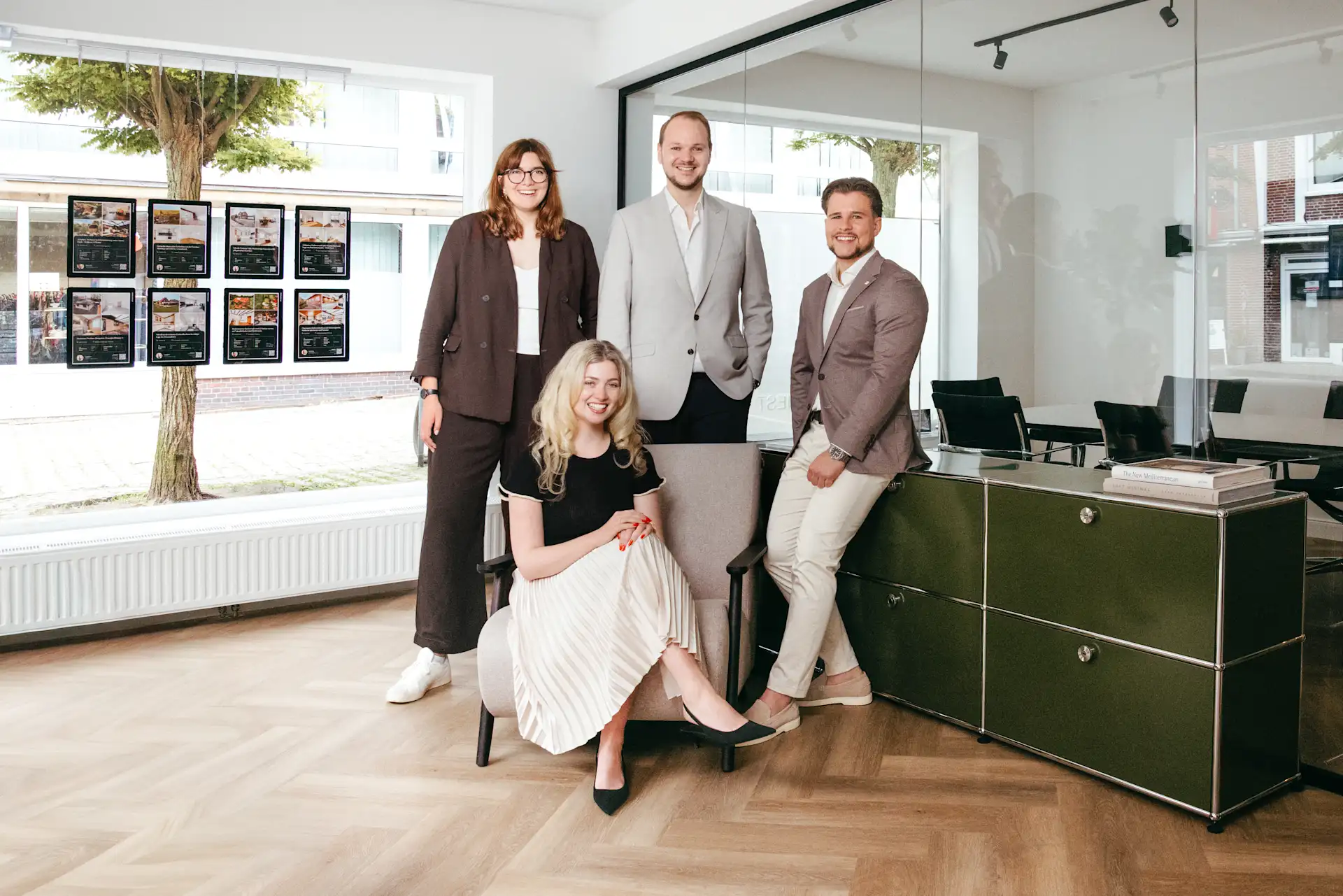 Vier Personen in Businesskleidung posieren in einem modernen Büro mit großen Fenstern und Holzboden.