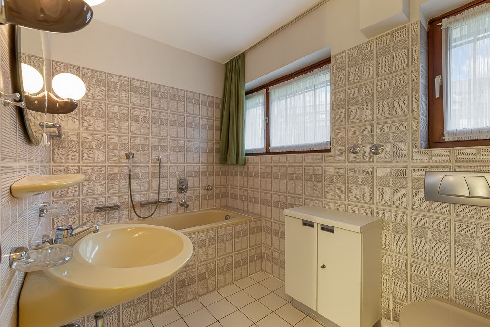 Badezimmer mit Badewanne, Waschbecken, Fliesenwänden und zwei Fenstern mit Vorhängen.