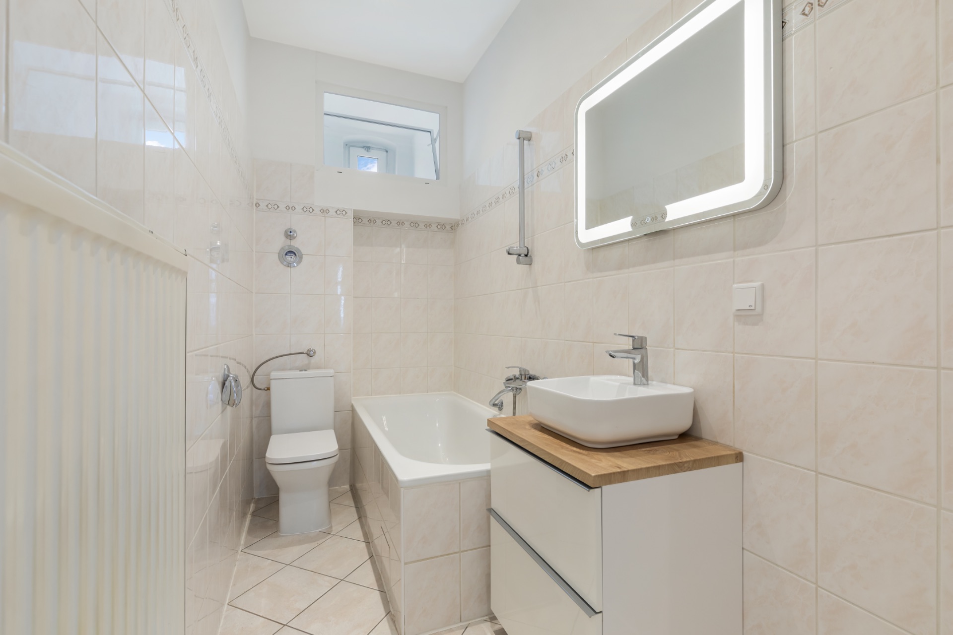 Helles Badezimmer mit Badewanne, Toilette, Waschbecken und beleuchtetem Spiegel.