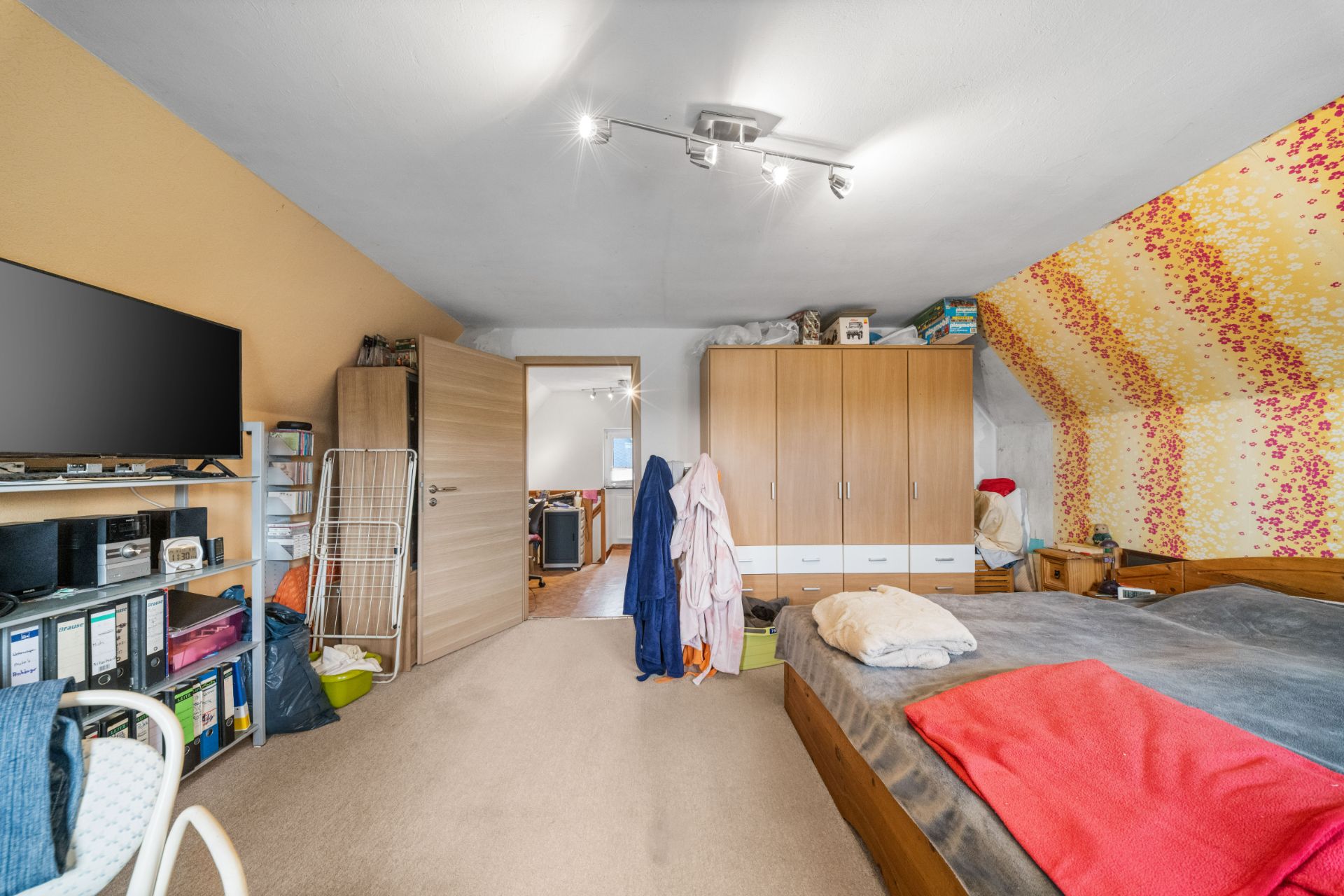 Schlafzimmer mit großem Bett, Kleiderschrank, Fernseher und bunter Tapete an der Schräge.