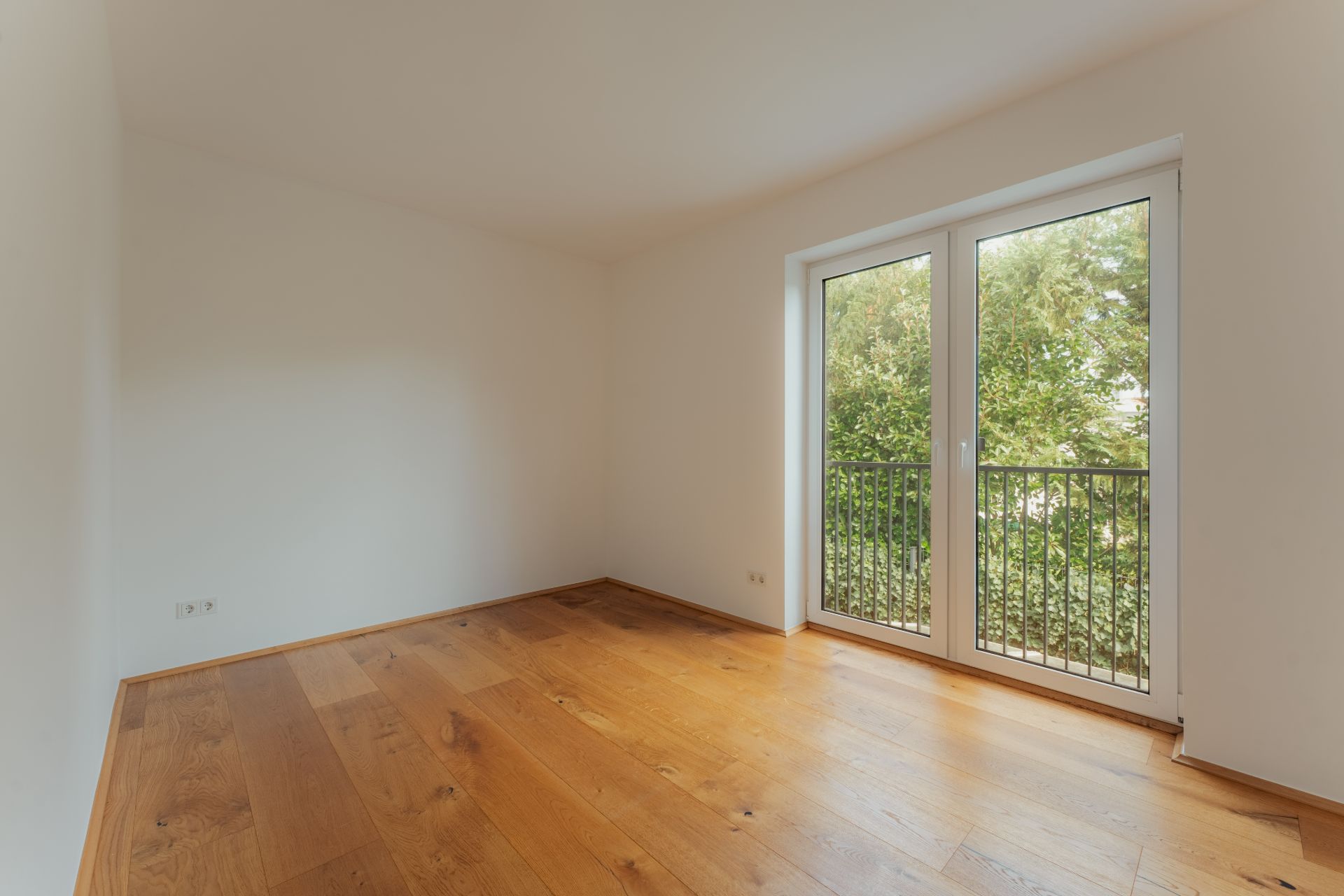 Leeres Zimmer mit Holzfußboden und bodentiefem Fenster, Blick auf Bäume.