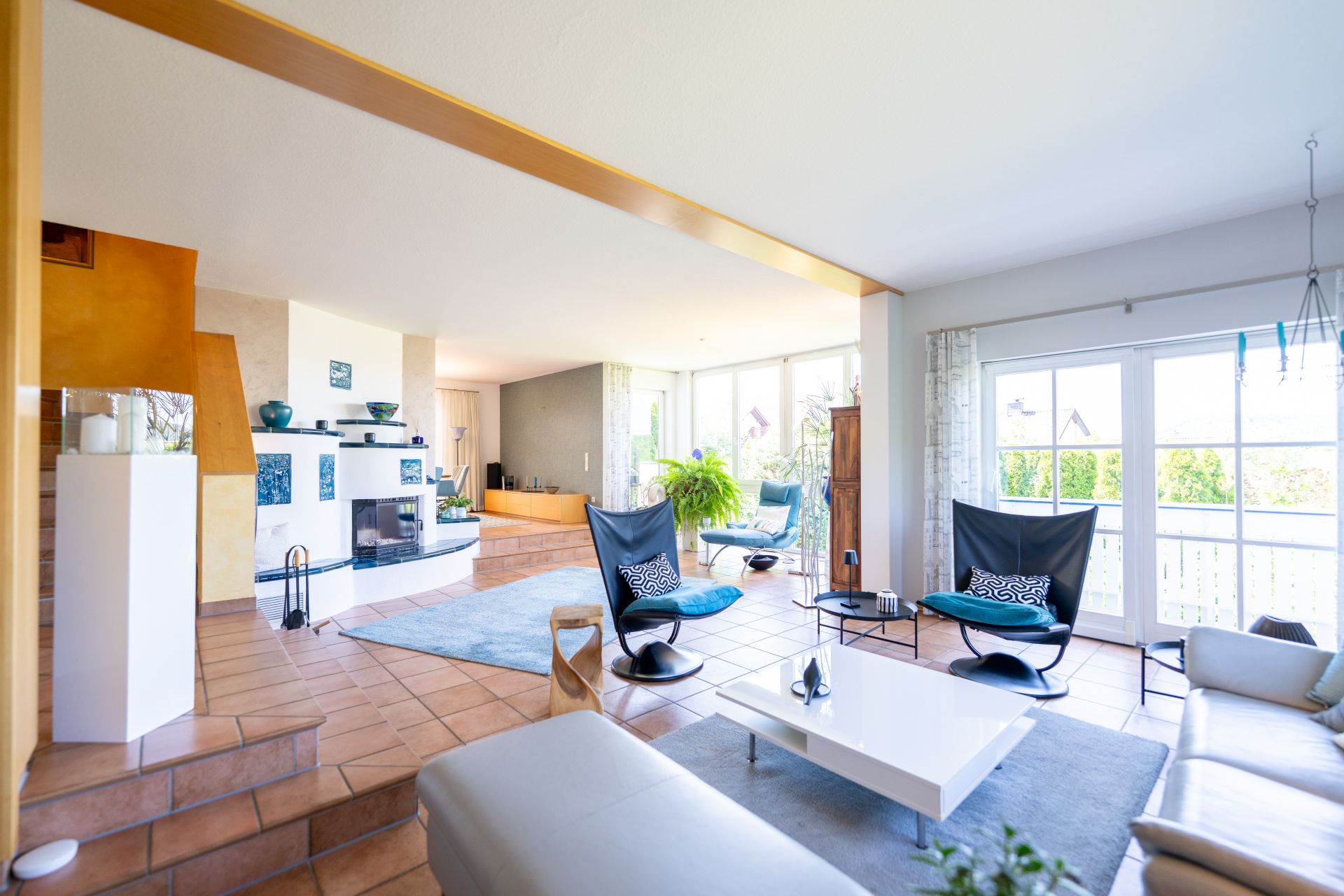 Modernes Wohnzimmer mit großen Fenstern, Sesseln, Sofa und weißem Couchtisch auf Fliesenboden.