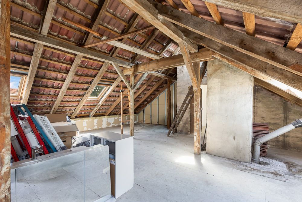 Dachboden mit sichtbaren Holzbalken, Dachziegeln, Fenstern und einigen gelagerten Gegenständen.
