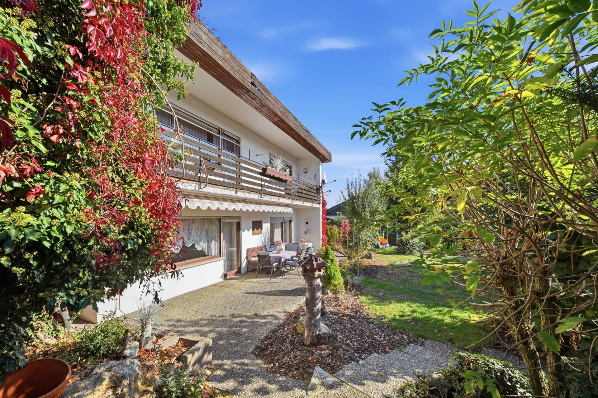 Zweistöckiges Wohnhaus mit Terrasse, Gartenmöbeln und Bewuchs, Blick auf den Garten bei sonnigem Wetter.
