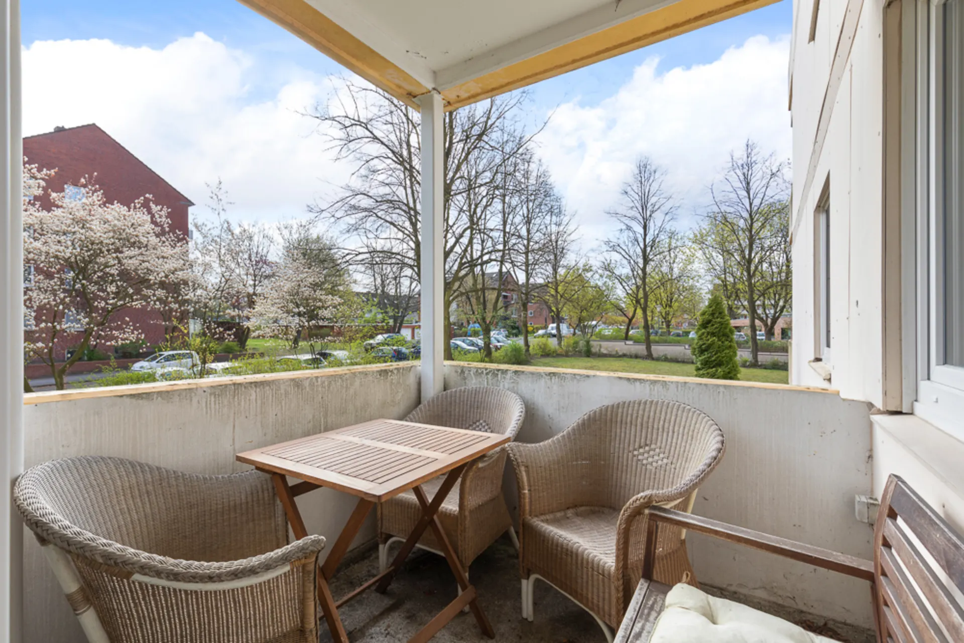 Balkon mit drei Korbstühlen und Holztisch, Blick auf Bäume und Wohnstraße.