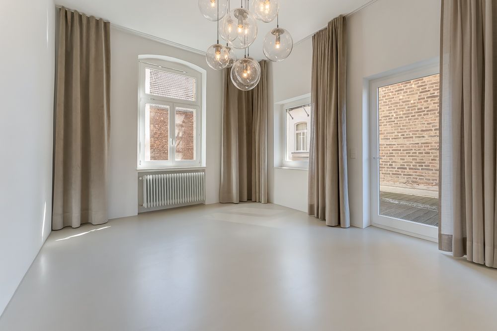 Leerer, heller Raum mit großen Fenstern, Vorhängen und moderner Deckenleuchte.