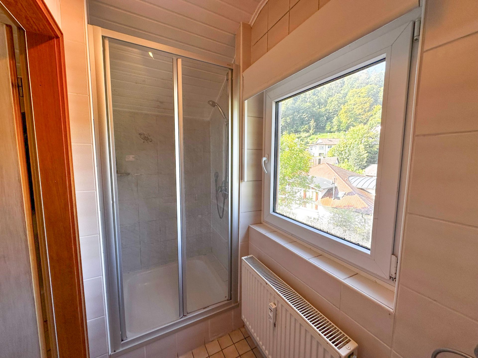 Dusche mit Glastür neben Fenster und Heizkörper in einem Badezimmer.