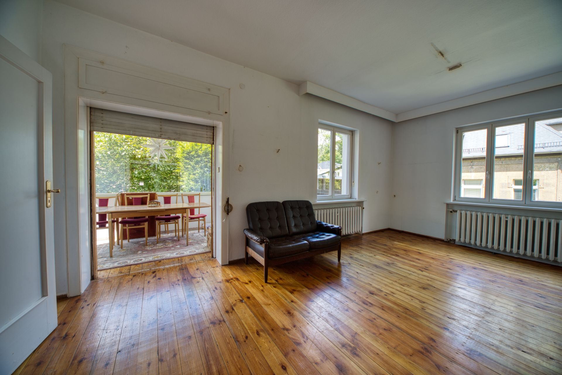 Leeres Wohnzimmer mit Holzboden, Sofa und Zugang zu einem verglasten Balkon mit Tisch und Stühlen.