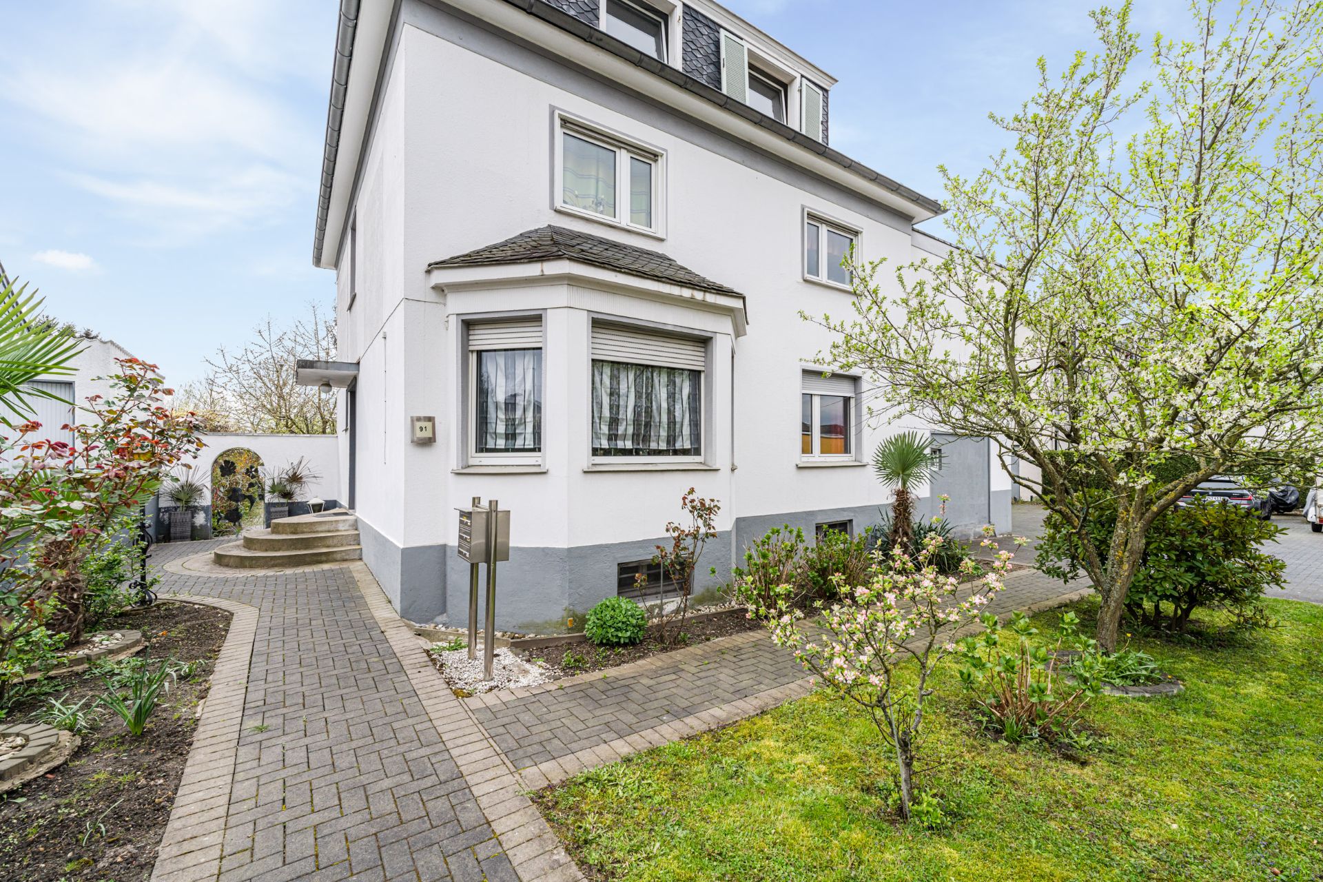 Weiße Wohnhausfassade mit Vorgarten, Bäumen und gepflastertem Weg.