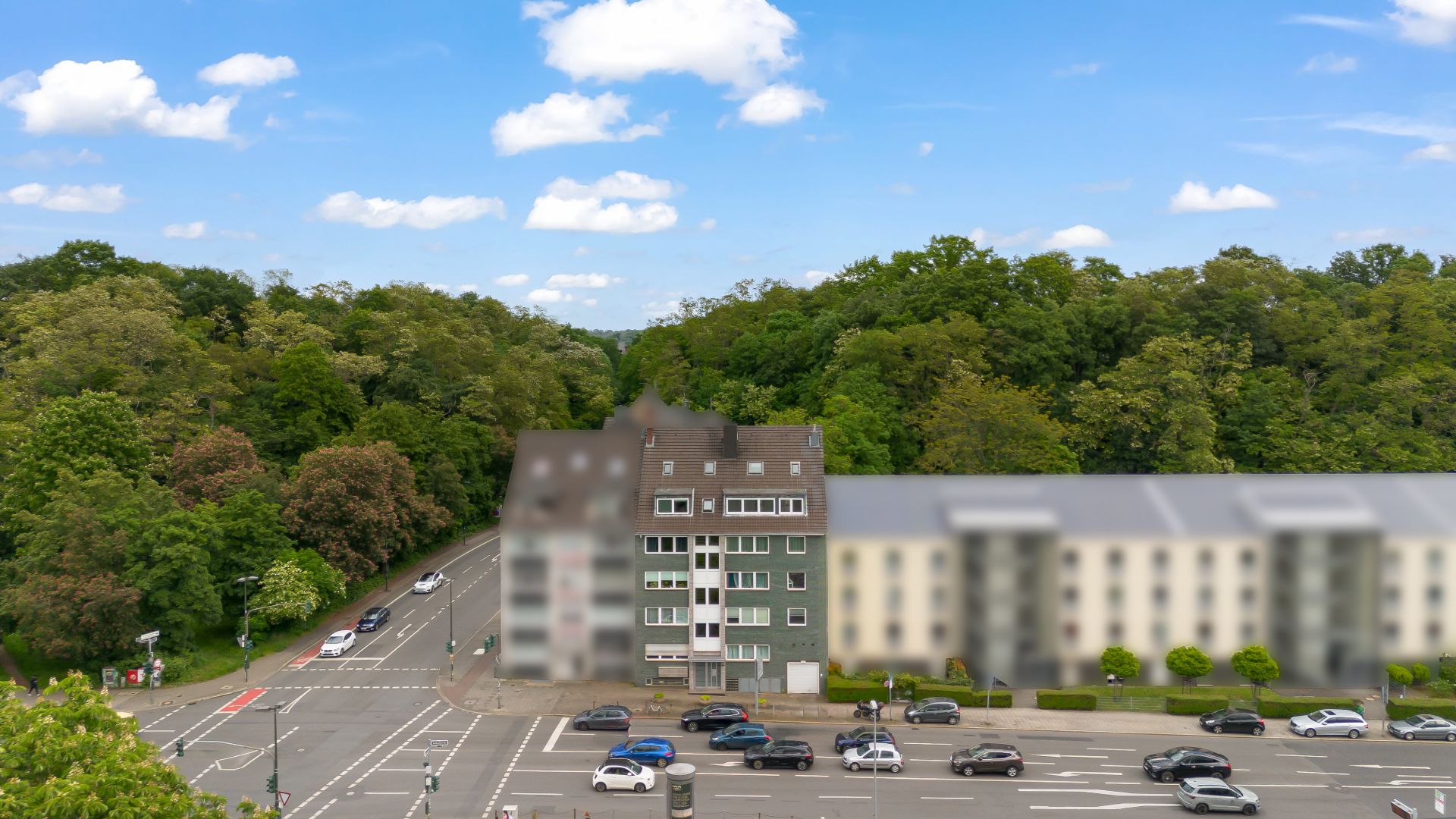 Mehrstöckiges Wohnhaus an einer Straßenkreuzung, im Hintergrund dichter grüner Wald.