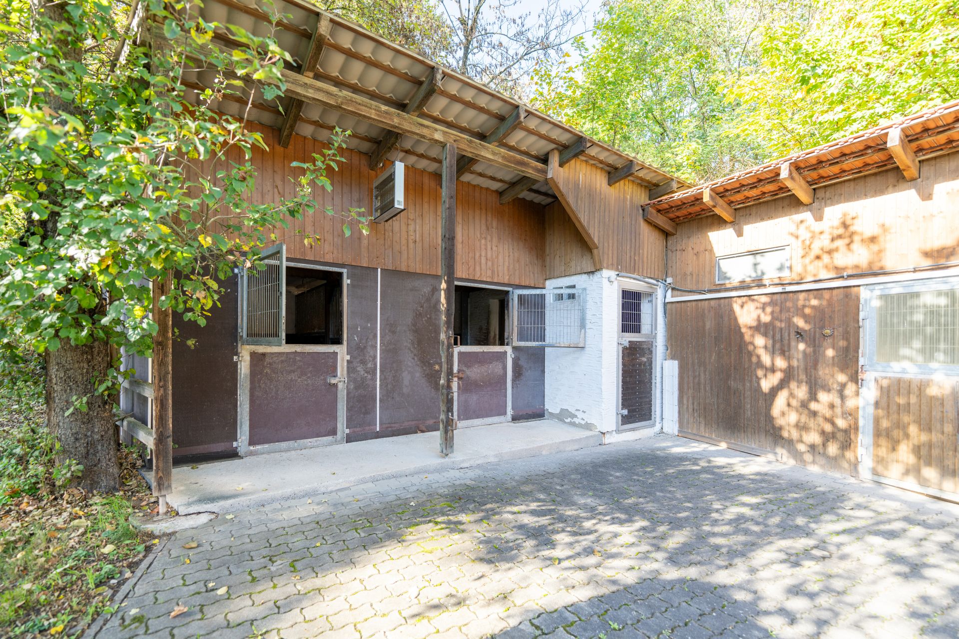 Offener Stall mit mehreren Boxen und gepflastertem Vorplatz, teils im Schatten von Bäumen.