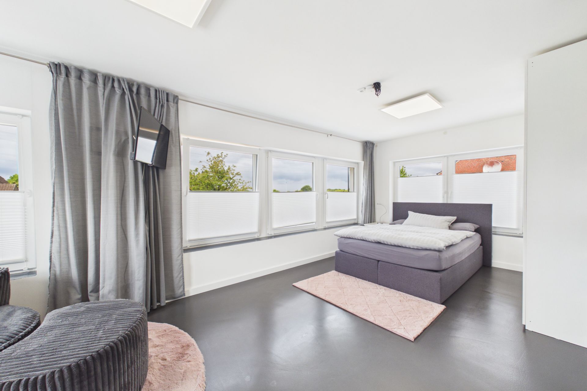 Helles Schlafzimmer mit großem Fenster, grauem Bett, Vorhängen und Teppich.