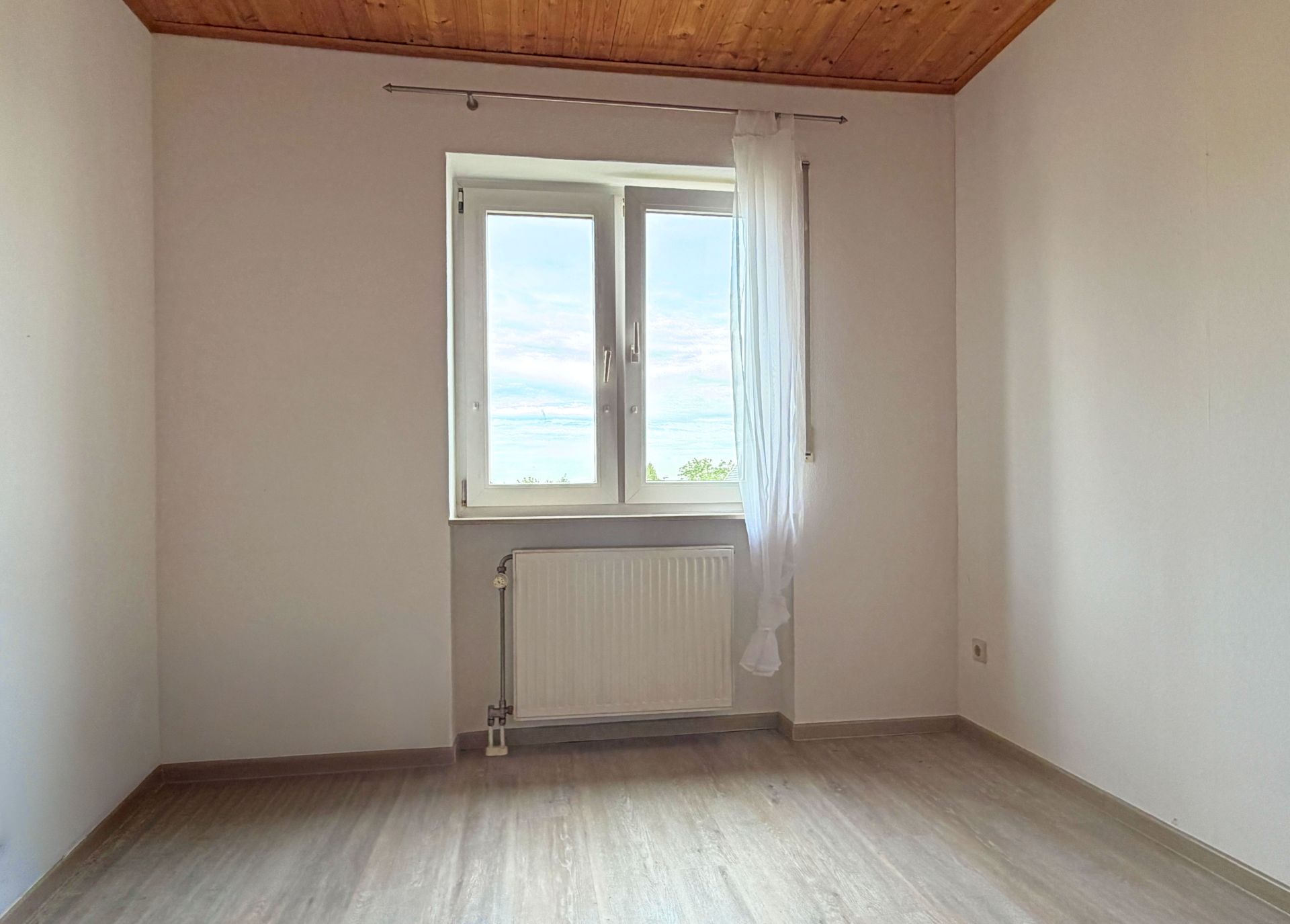Leeres Zimmer mit weißer Wand, Holzdecke, Fenster mit Vorhang und Heizkörper darunter.