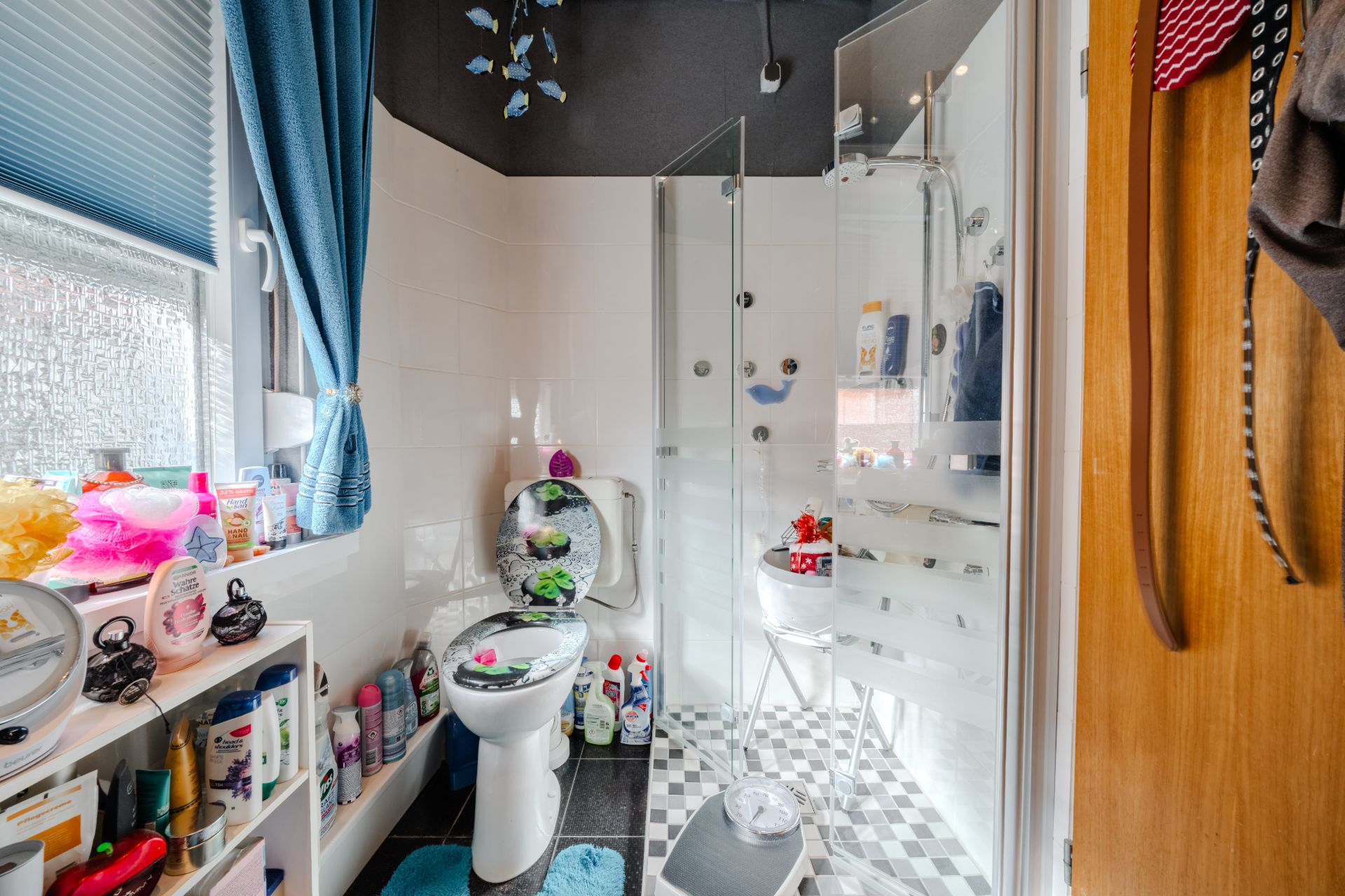 Kleines Badezimmer mit Toilette, Dusche, Fenster und Regal mit Pflegeprodukten.