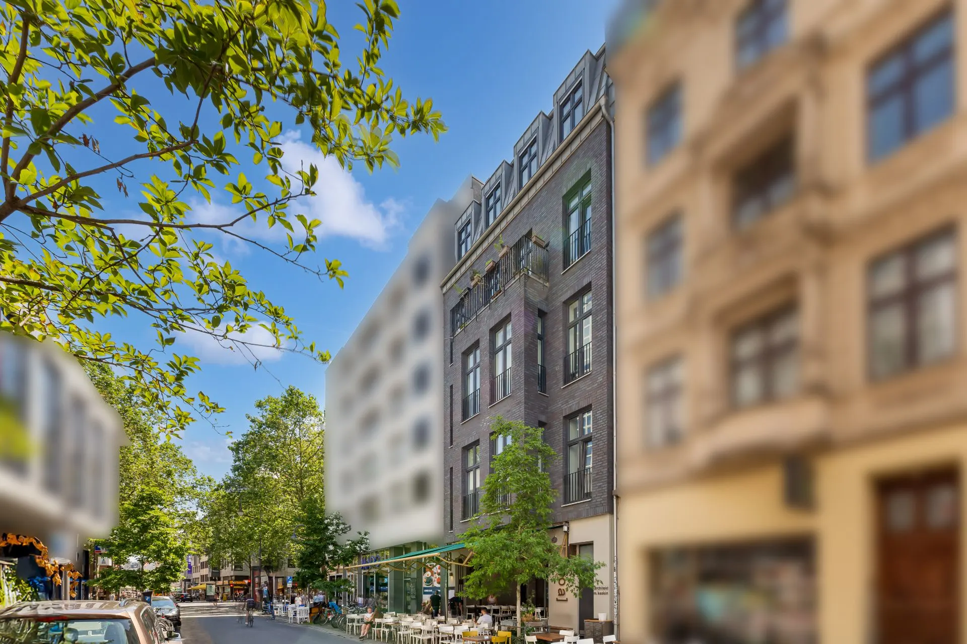 Mehrstöckiges Wohnhaus mit großen Fenstern und Außencafé an einer belebten Straße mit Bäumen.