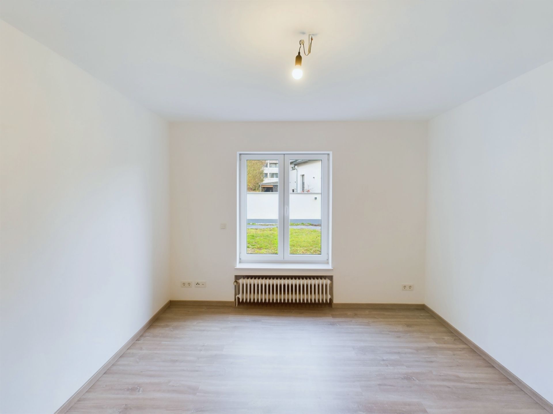 Leerer Raum mit Holzboden, weißer Wand, Fenster und Heizkörper darunter.