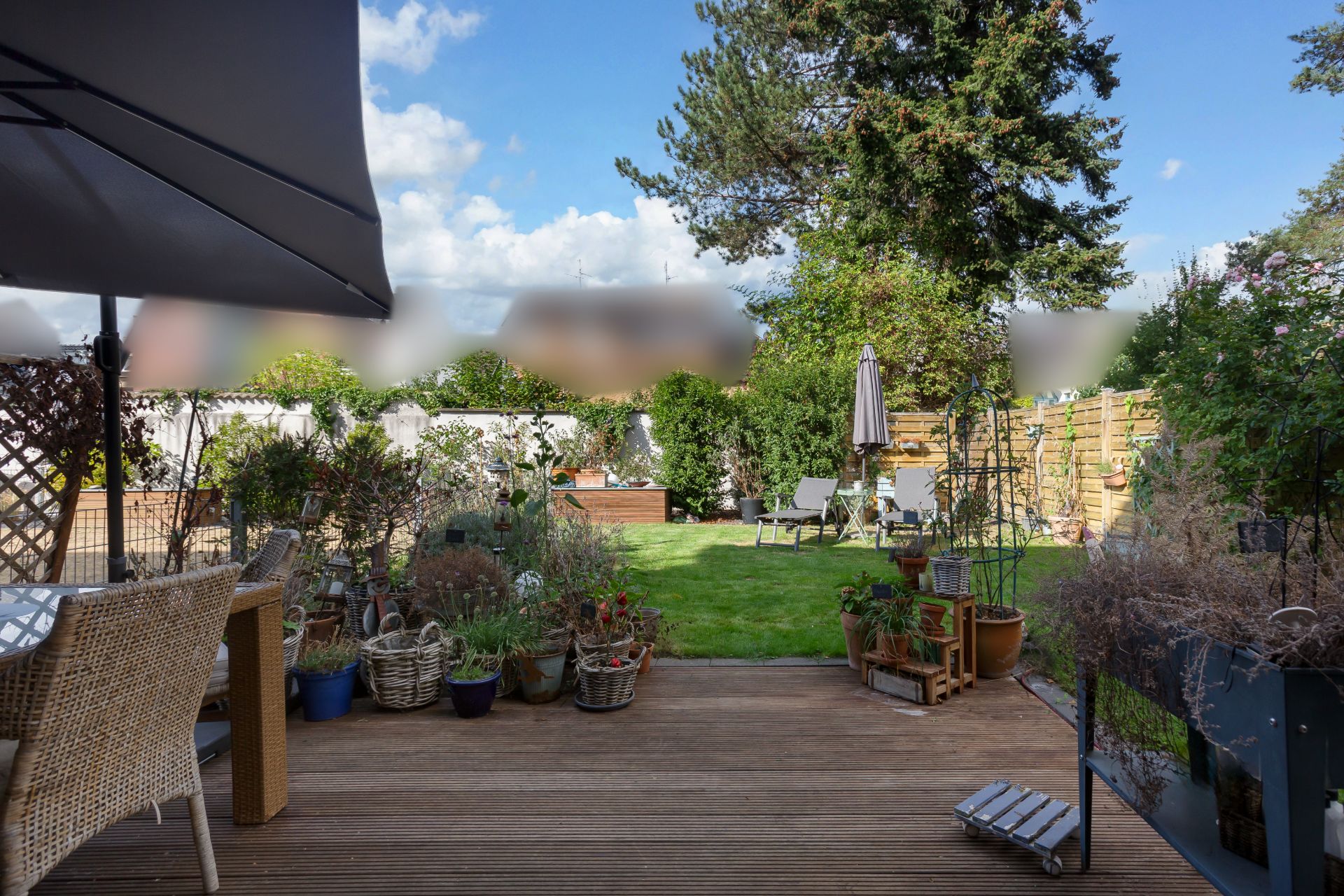 Terrasse mit Holzboden, vielen Pflanzen in Töpfen und Blick auf einen grünen Garten mit Liegestühlen.