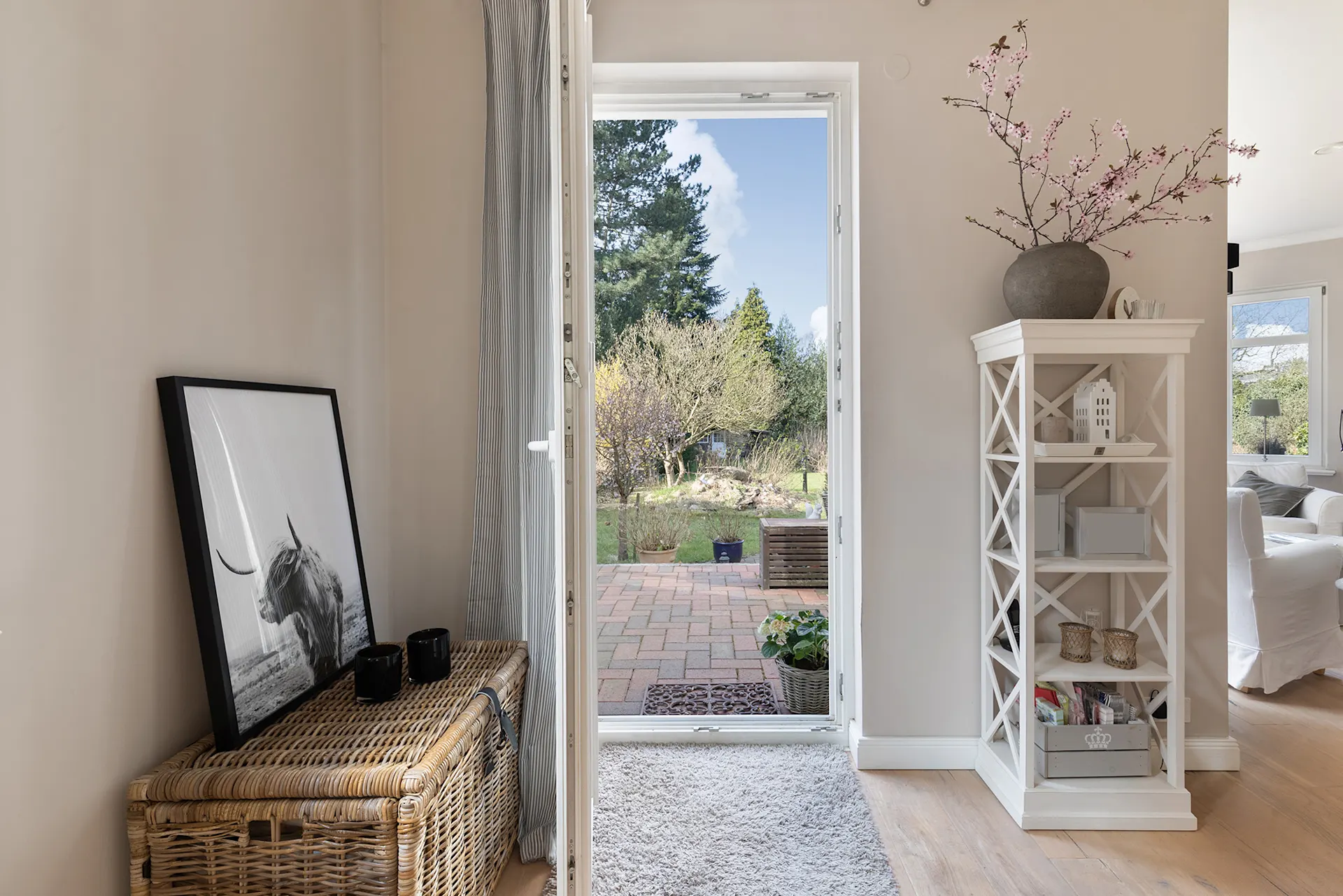 Offene Tür zum Garten, Korb mit Bild, weißes Regal und blühende Zweige im Eingangsbereich.