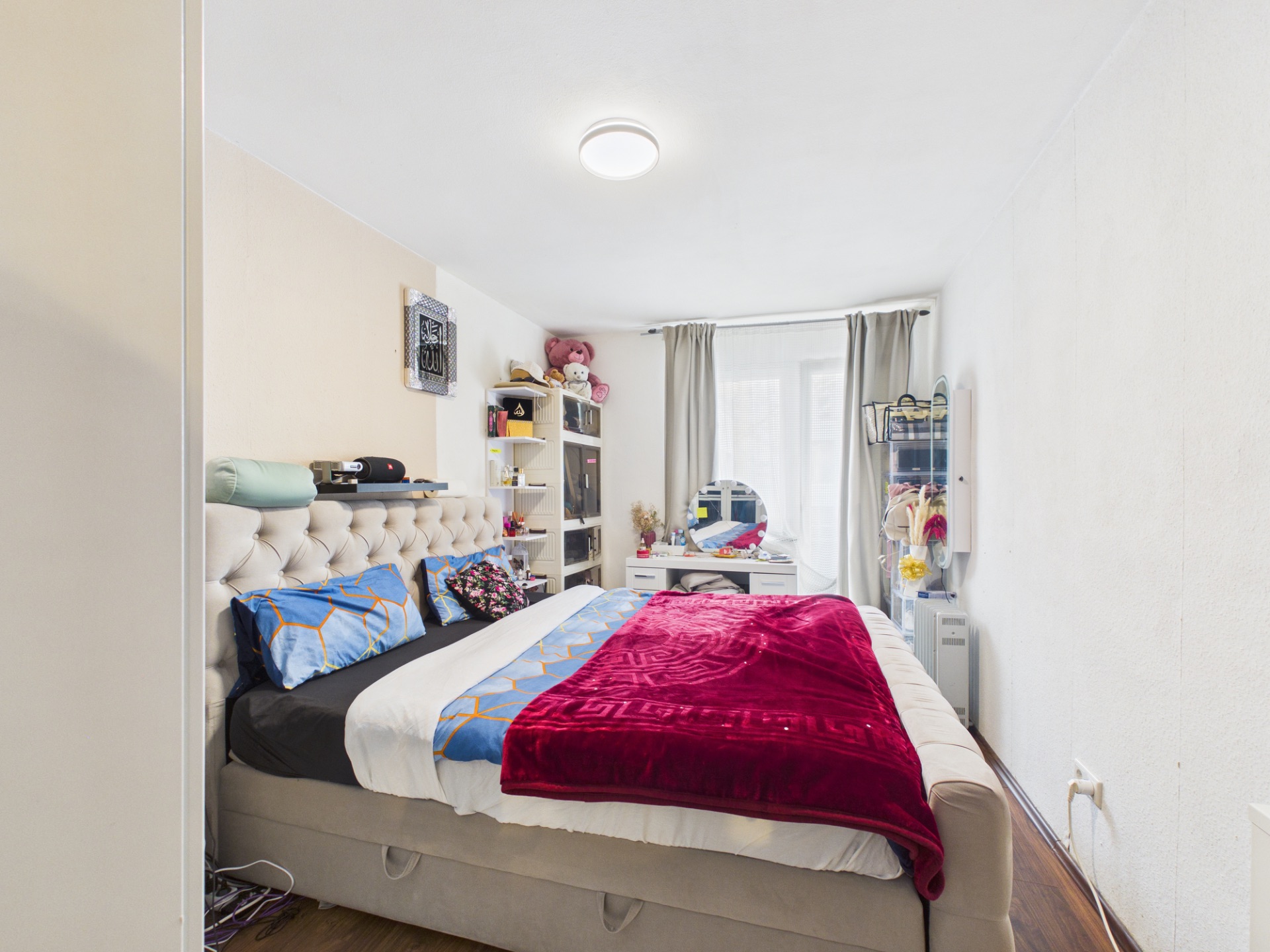 Schlafzimmer mit großem Bett, Regal, Schminktisch und Fenstern mit grauen Vorhängen.