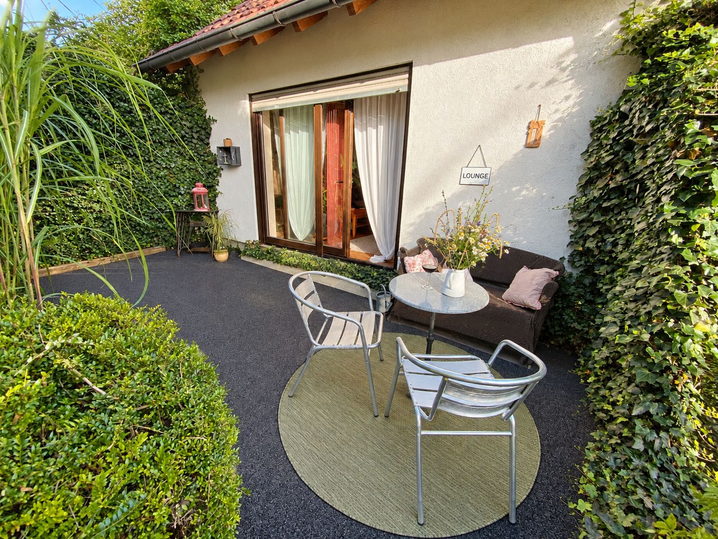 Kleine Terrasse mit zwei Stühlen, Tisch, Sofa und Pflanzen vor einer Glastür.
