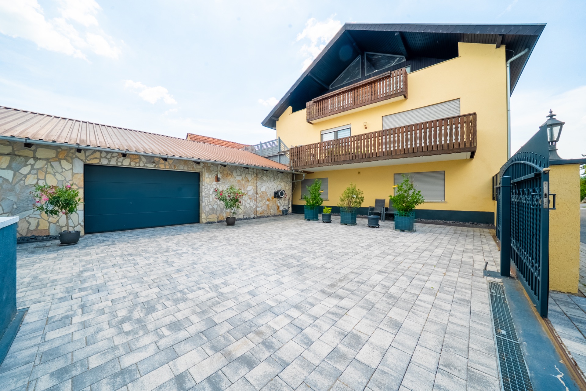Gepflasterter Hof mit gelbem Wohnhaus, Balkon und Garage, mehrere Blumenkübel am Rand.