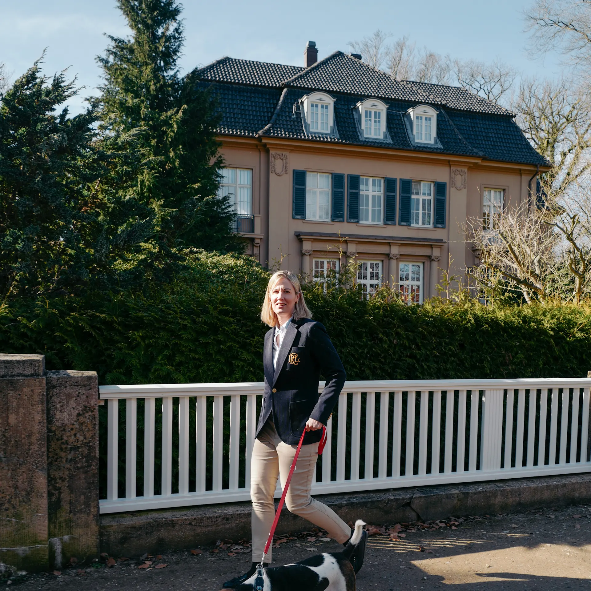 Frau geht mit Hund an einem weißen Zaun vor einem großen Wohnhaus spazieren.
