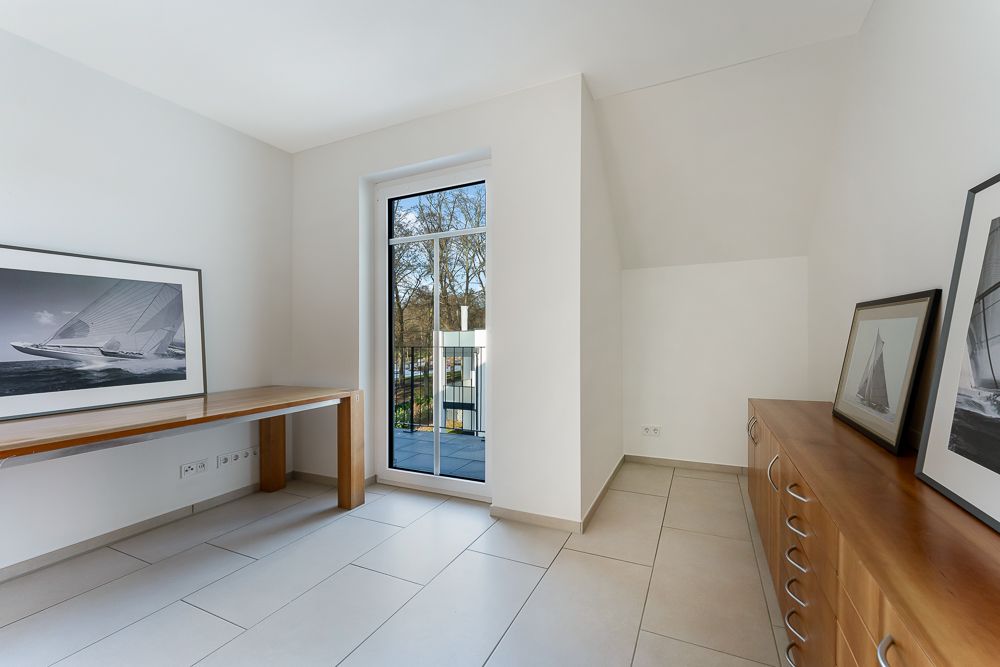 Helles Zimmer mit Fliesenboden, Schreibtisch, Kommode und großen Fenstern, Bilder an der Wand.