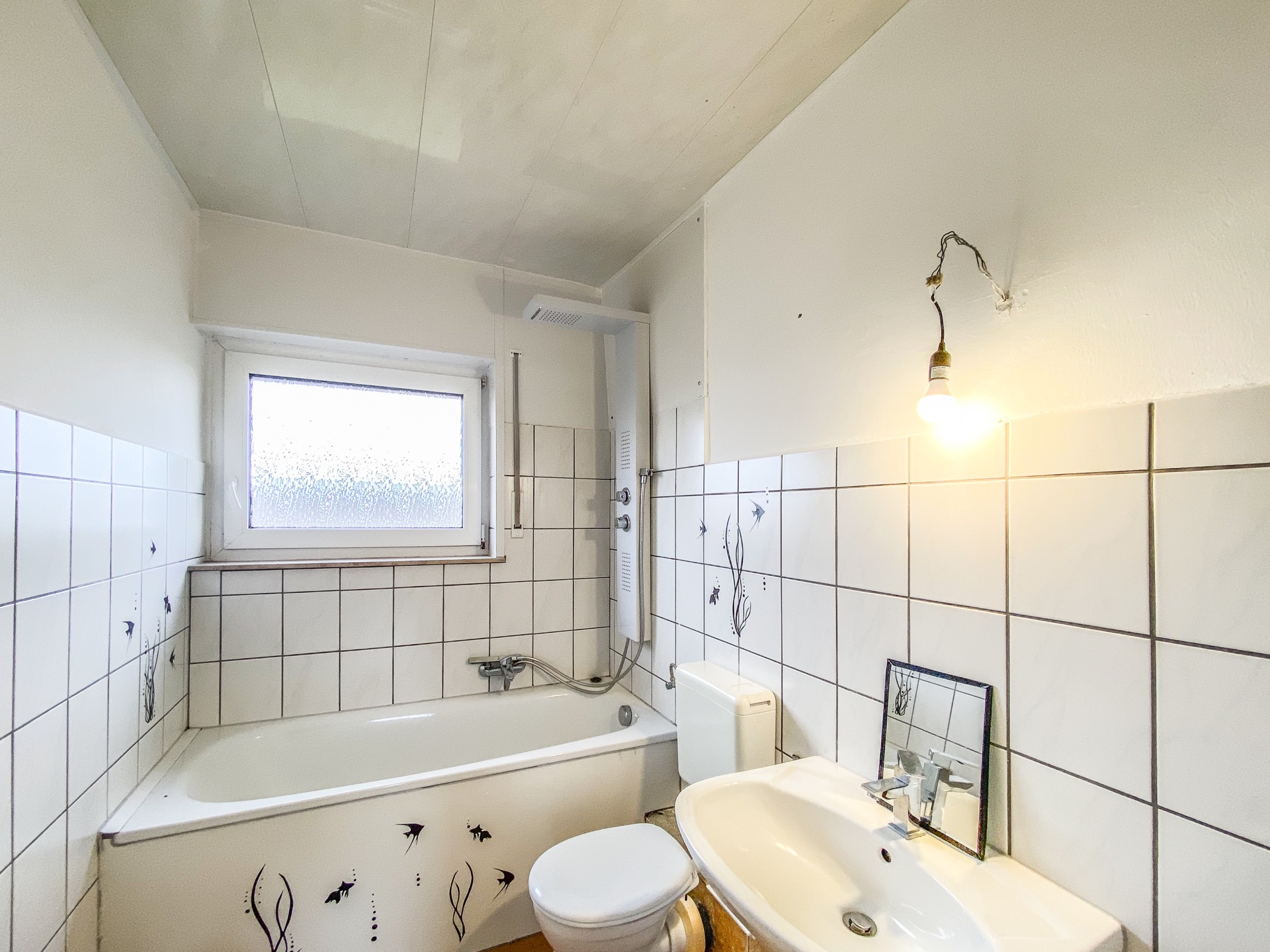 Helles Badezimmer mit Badewanne, Waschbecken, Toilette und Fenster, weiße Fliesen mit Dekor.
