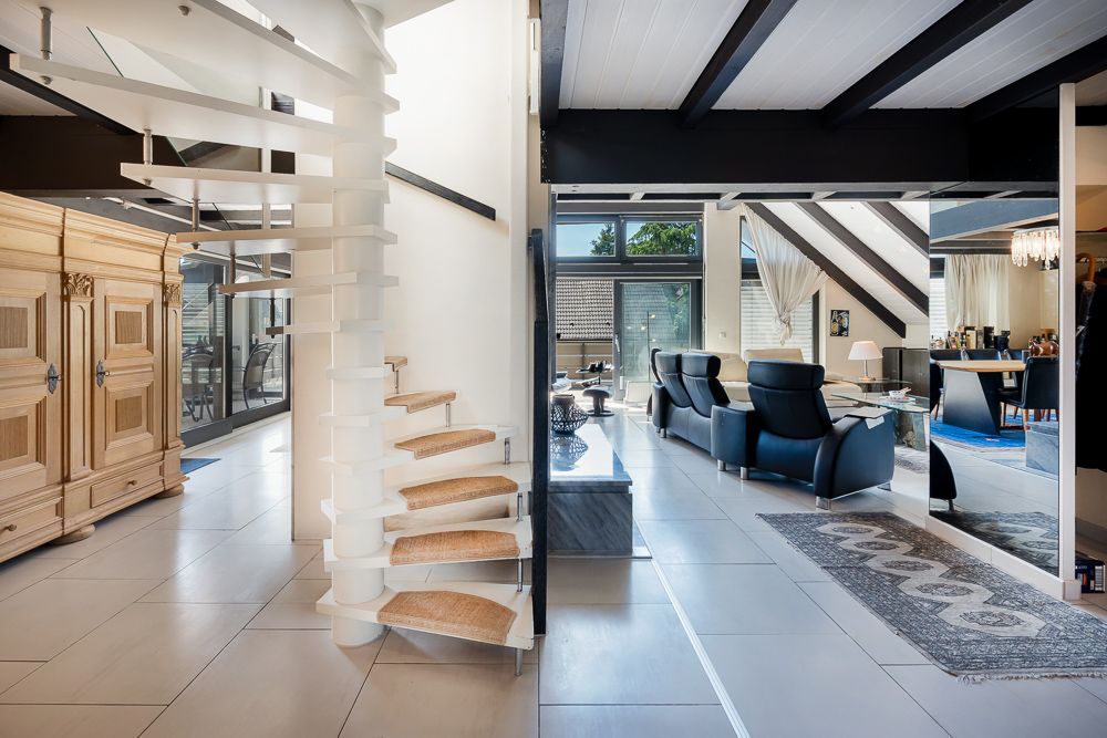 Helles Wohnzimmer mit moderner Wendeltreppe und großen Fenstern, zwei schwarzen Sesseln und Teppich.