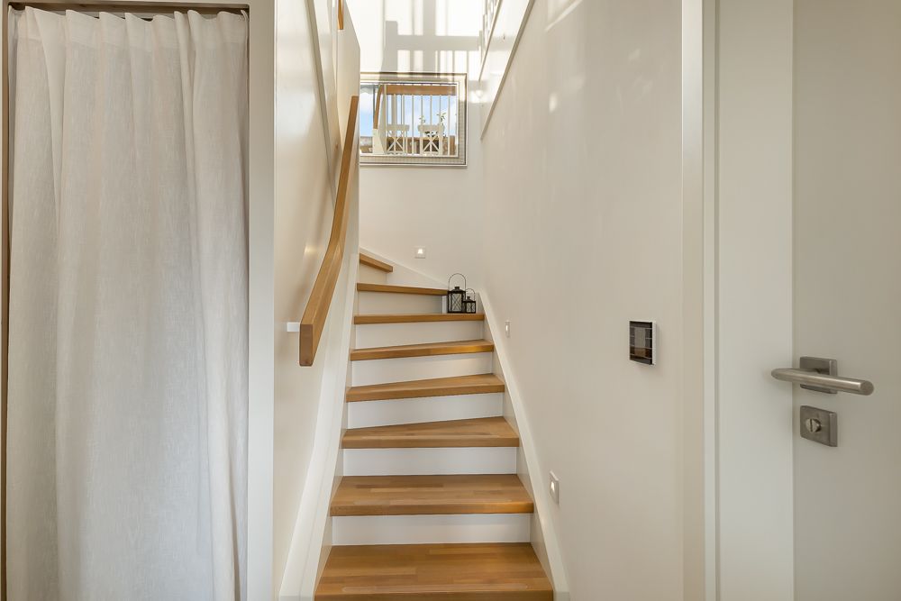 Helle Holztreppe mit Geländer, Fenster im Hintergrund, Vorhang links, weiße Wände.