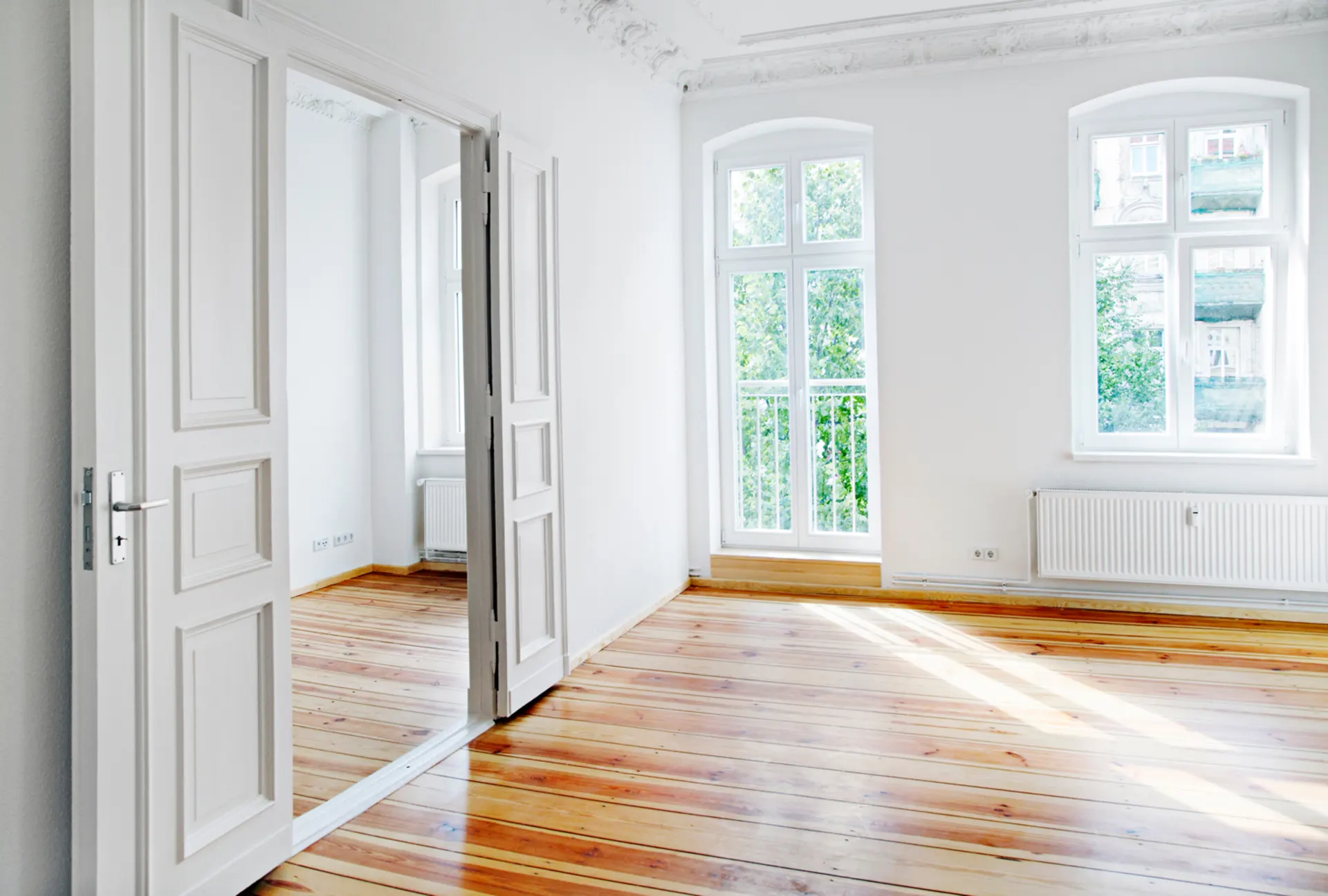 Helles, leeres Zimmer mit Holzboden, weißen Wänden und großen Fenstern.
