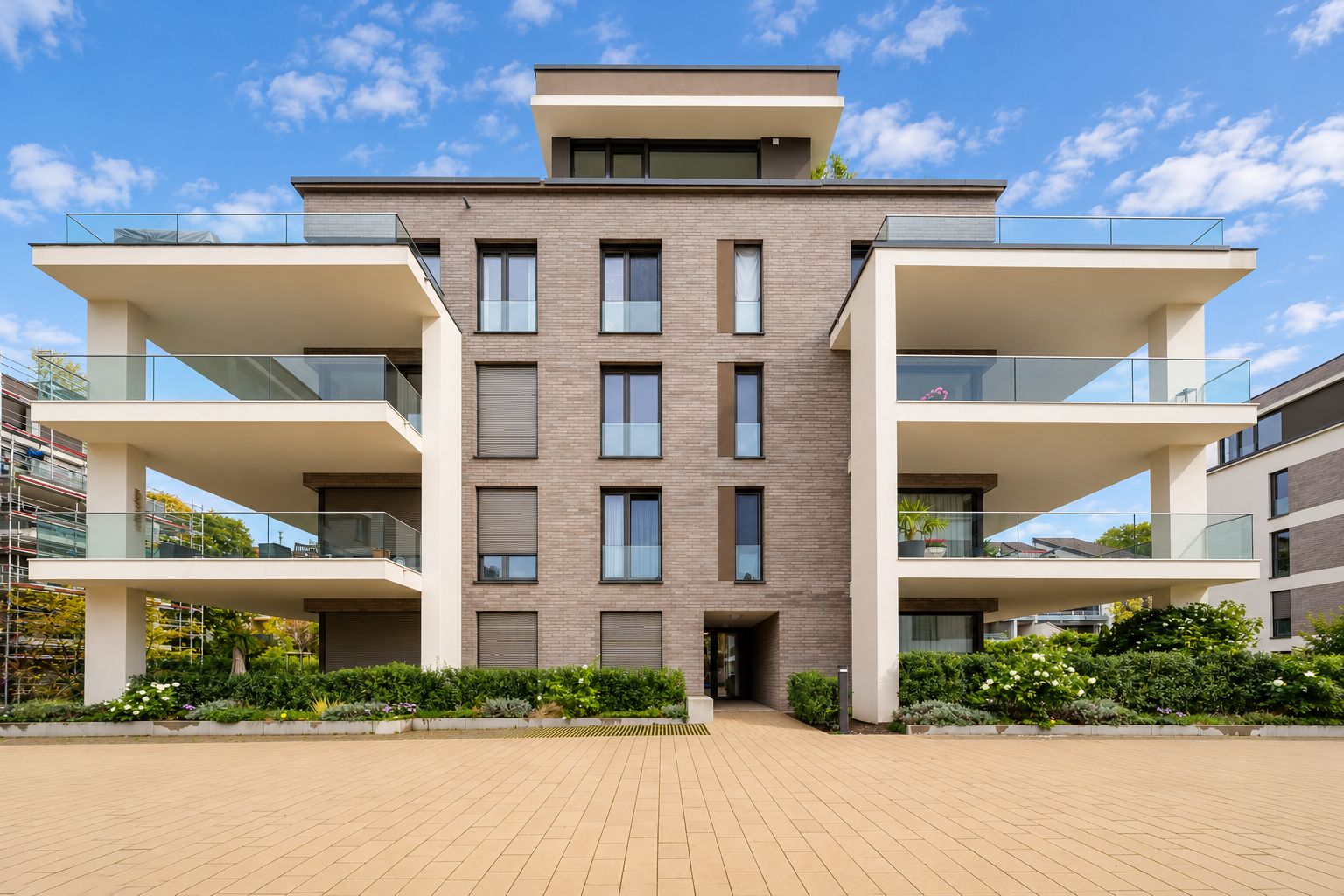 Modernes Wohngebäude mit großen Balkonen und Glasgeländern, davor ein gepflasterter Vorplatz.