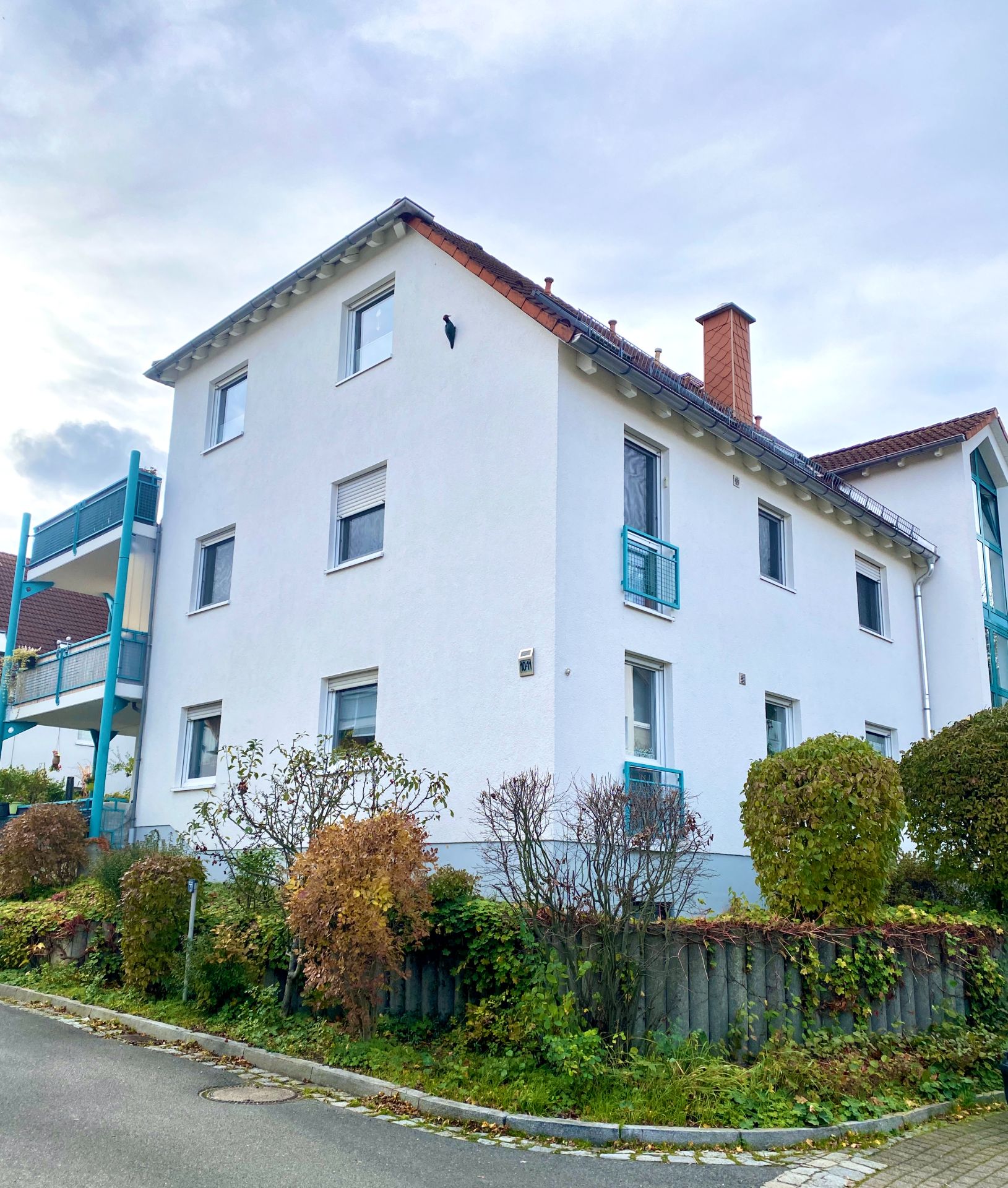 Weißes Wohnhaus mit rotem Dach und mehreren Fenstern, umgeben von Sträuchern.