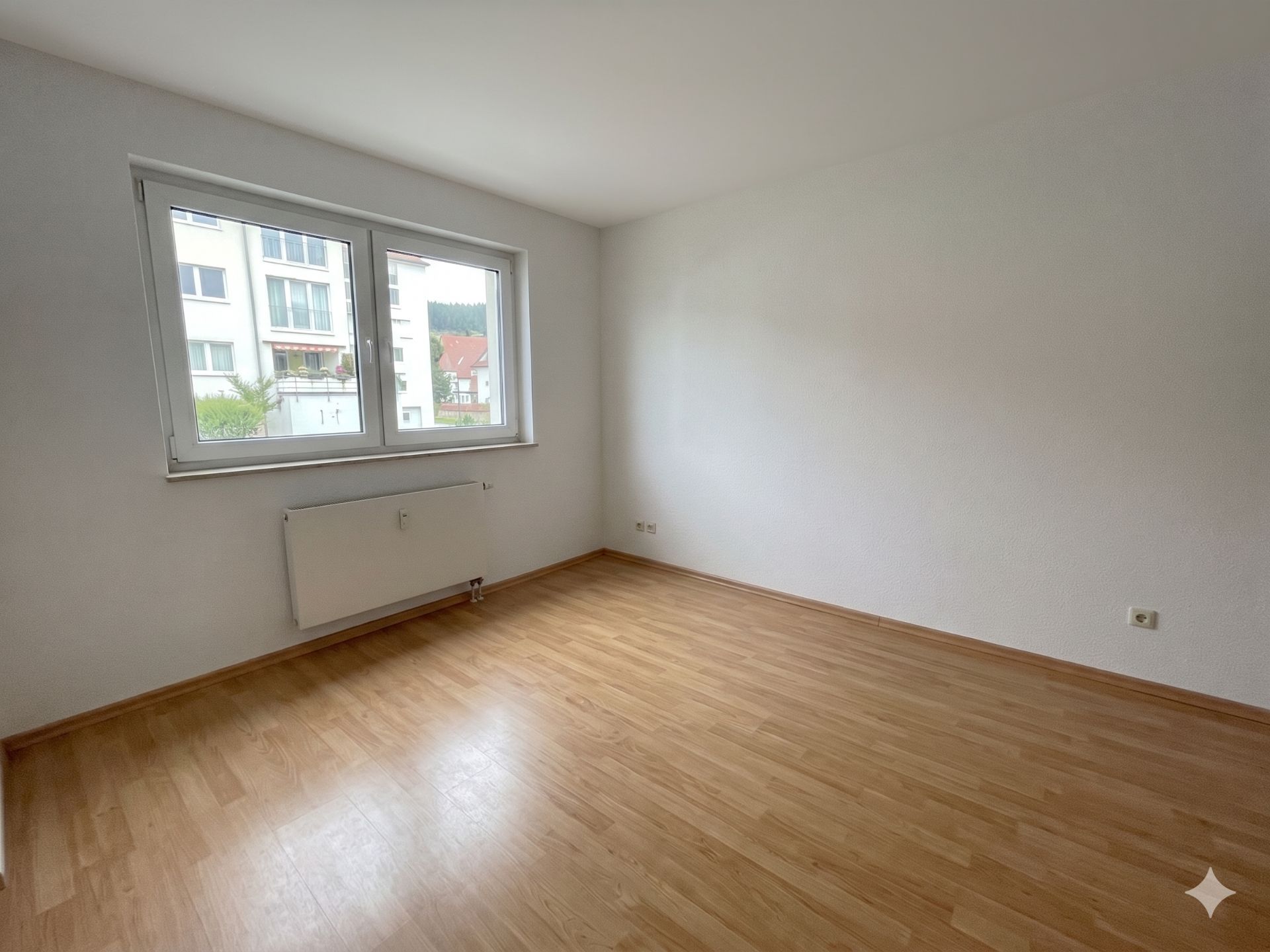 Leeres Zimmer mit hellem Laminatboden, weißer Wand und großem Fenster.