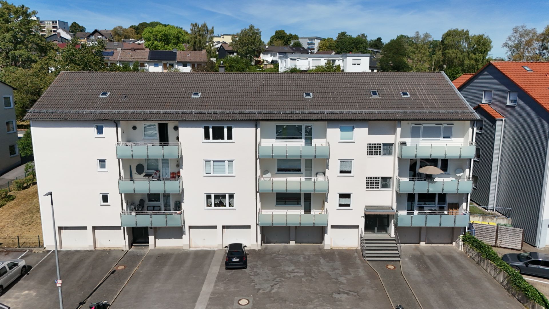 Dreistöckiges Wohnhaus mit Balkonen, Garagen und leerem Parkplatz davor.