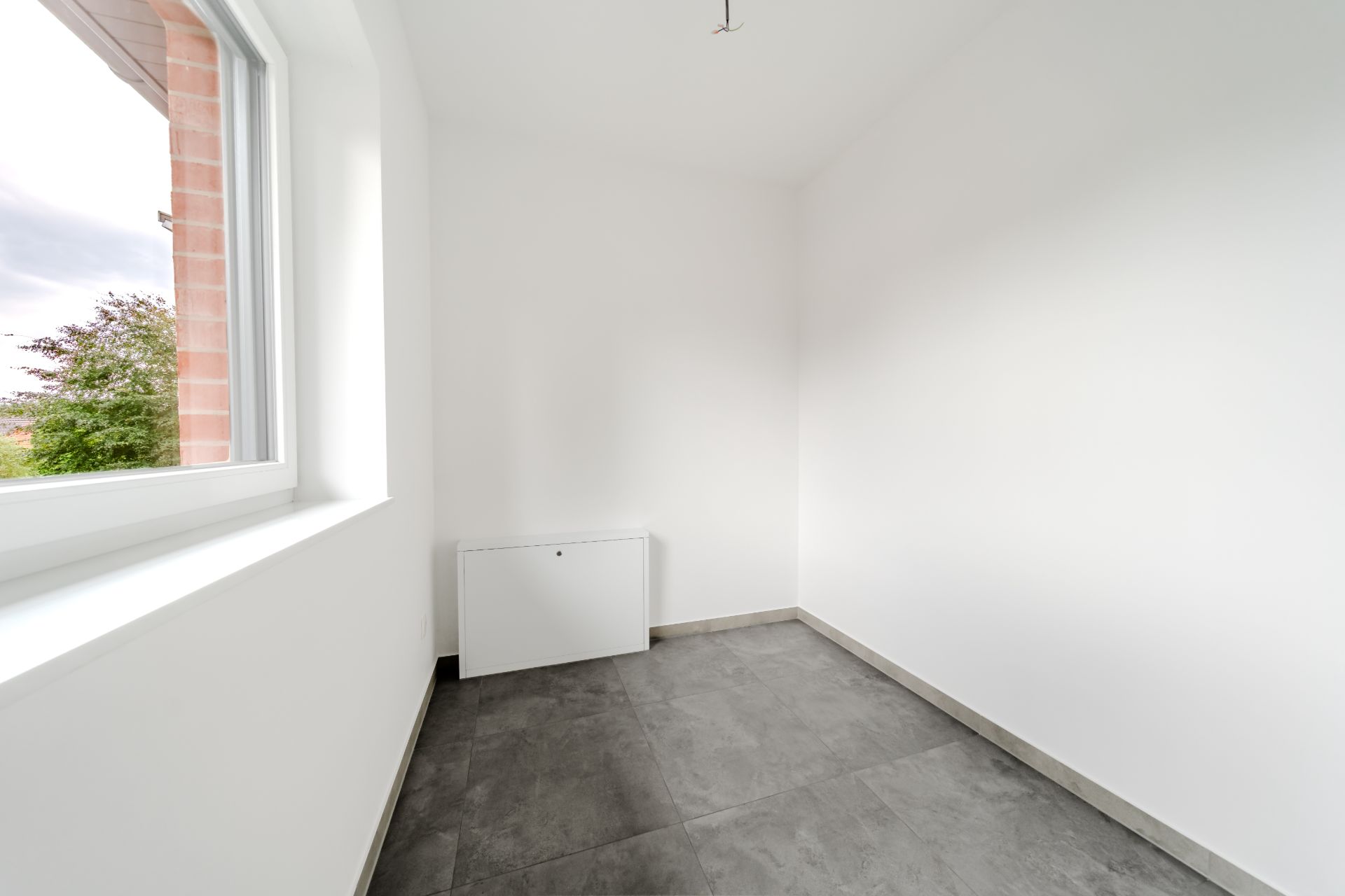Heller leerer Raum mit Fenster, weißen Wänden und grauem Fliesenboden.