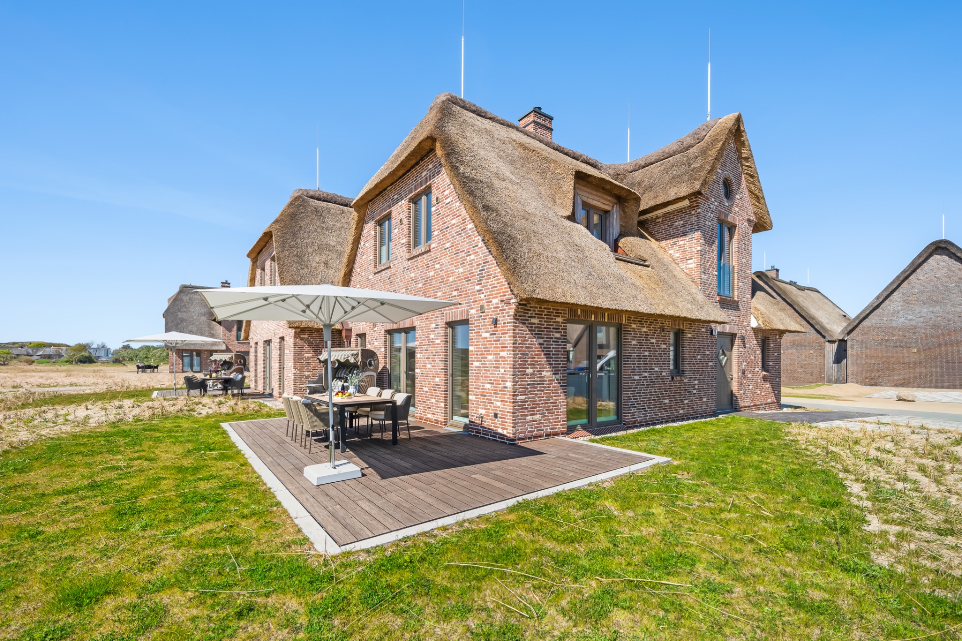 Backsteinhaus mit Reetdach, Terrasse mit Esstisch und Sonnenschirm auf einer Wiese.