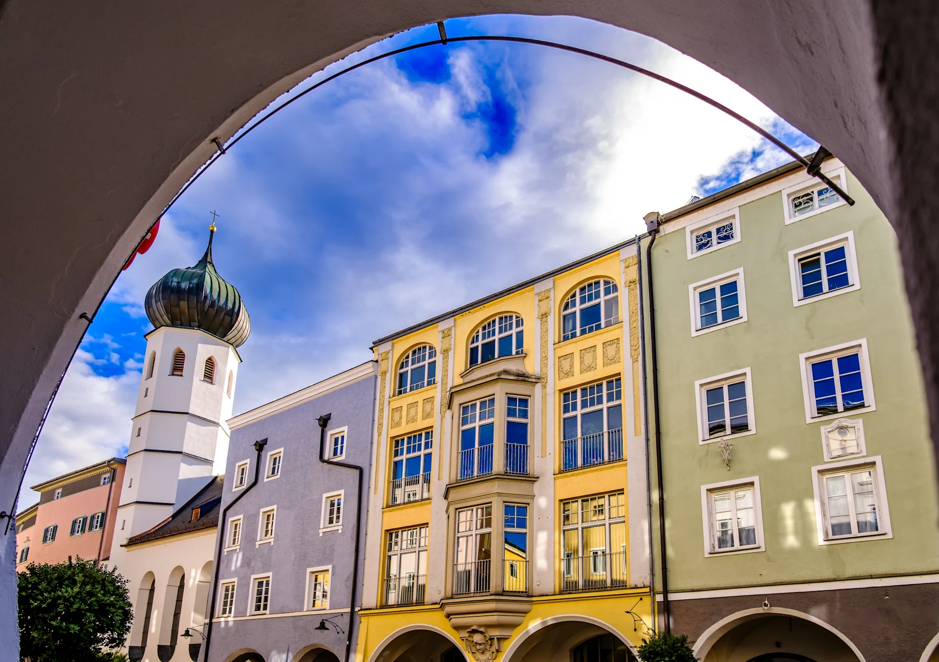 Bunte Wohngebäude und Kirchturm hinter einem Torbogen bei blauem Himmel.