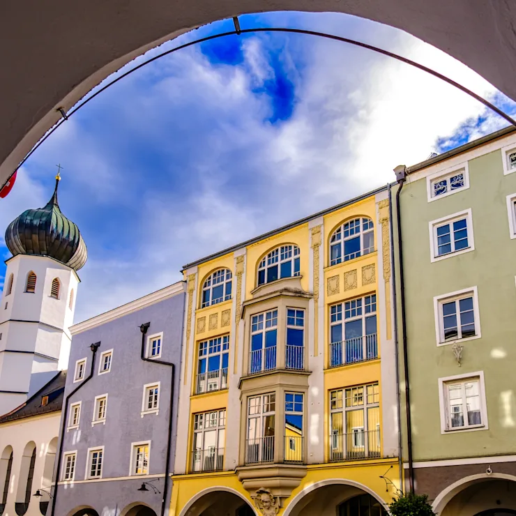 Bunte Wohngebäude und Kirchturm hinter einem Torbogen bei blauem Himmel.