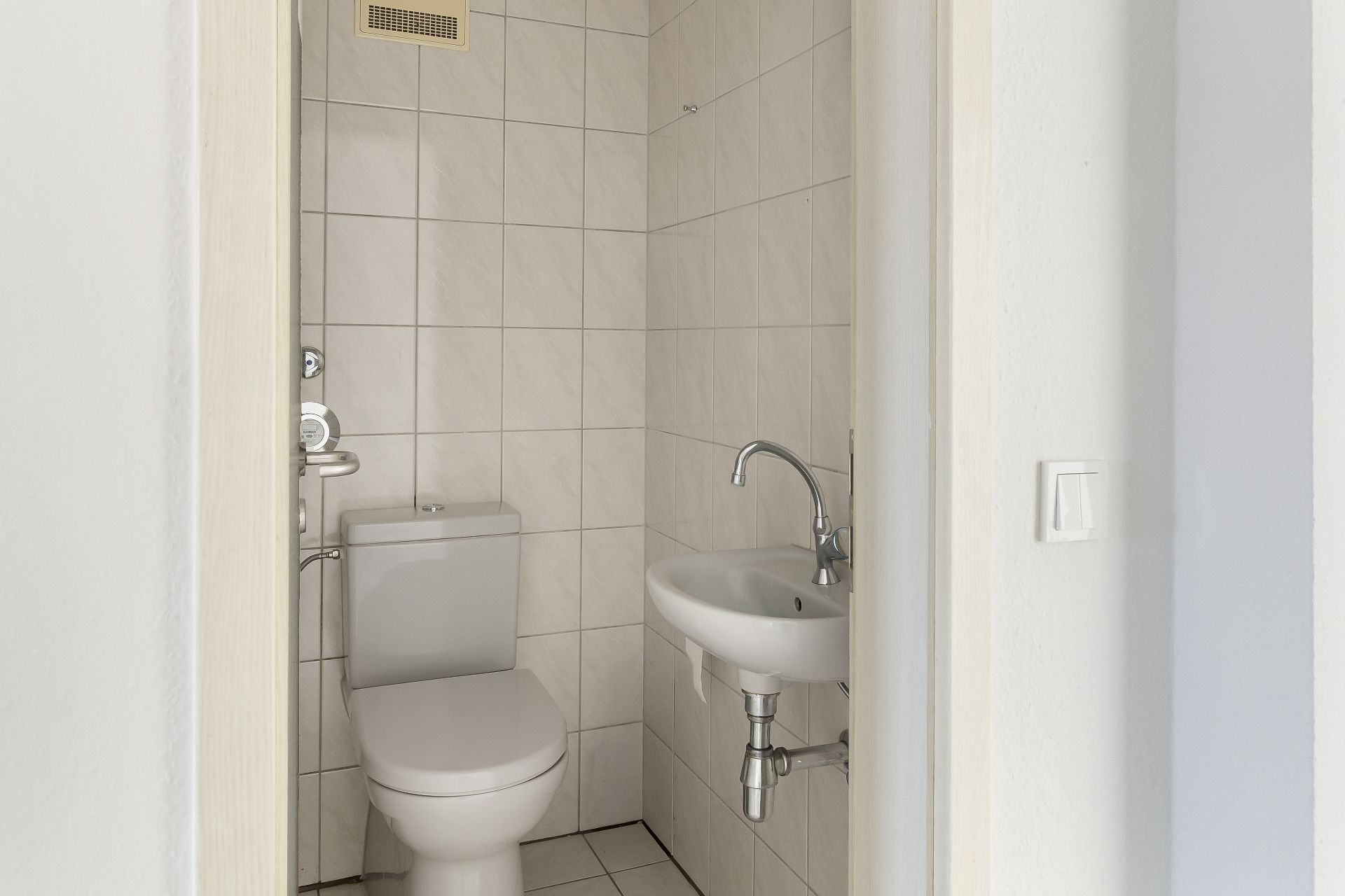 Kleines Badezimmer mit WC und Wandwaschbecken, weiße Fliesen an Wänden und Boden.
