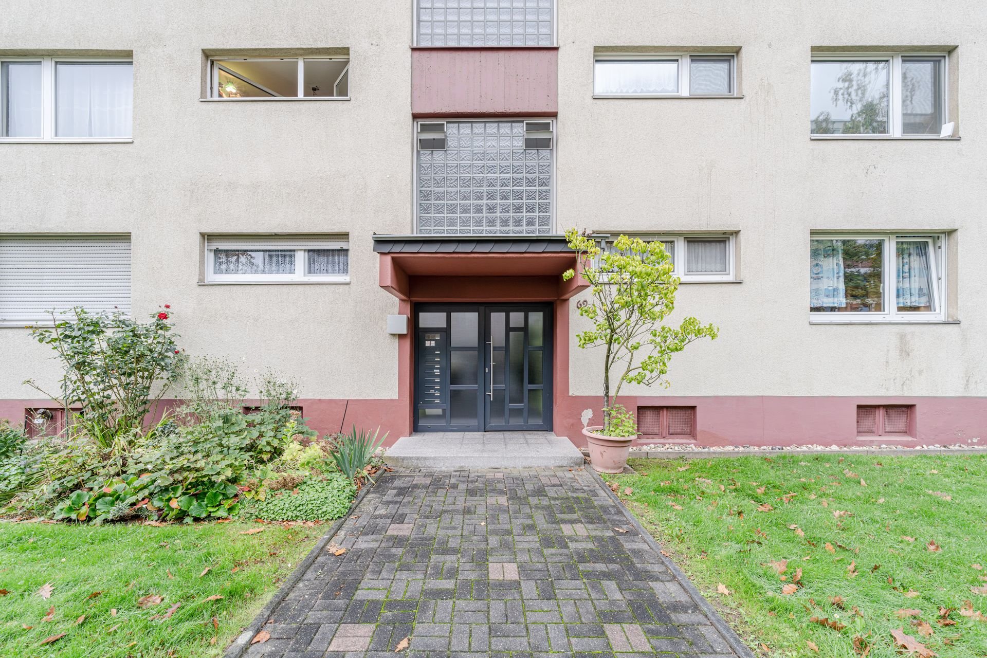 Eingang eines Mehrfamilienhauses mit Glasdach, Pflasterweg und kleiner Grünfläche davor.