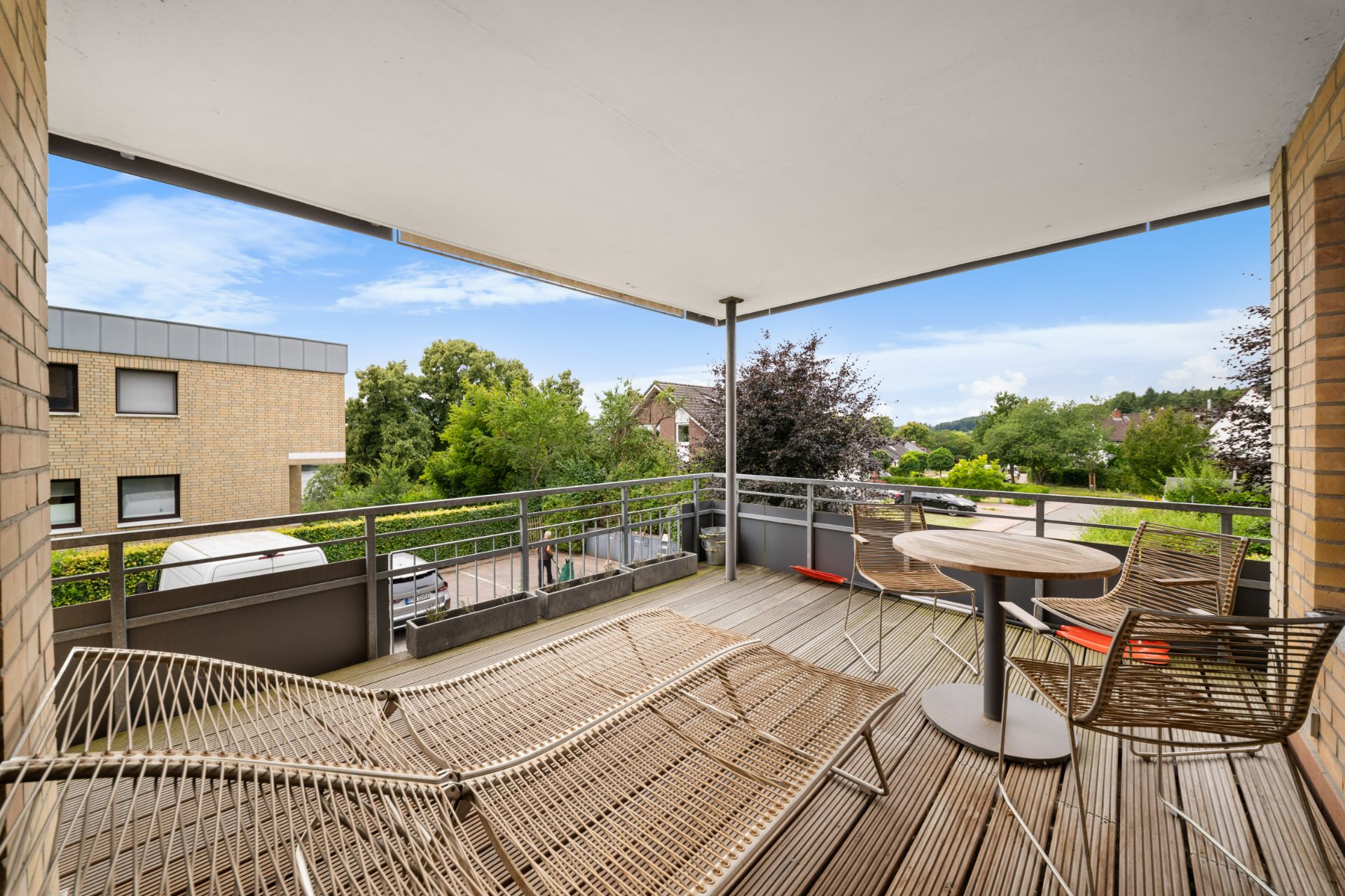 Überdachter Balkon mit zwei Liegen, einem runden Tisch und zwei Stühlen, Blick ins Grüne.