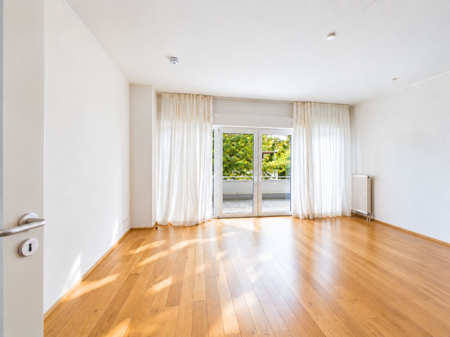 Heller, leerer Raum mit Holzboden, großen Fenstern und weißen Vorhängen.