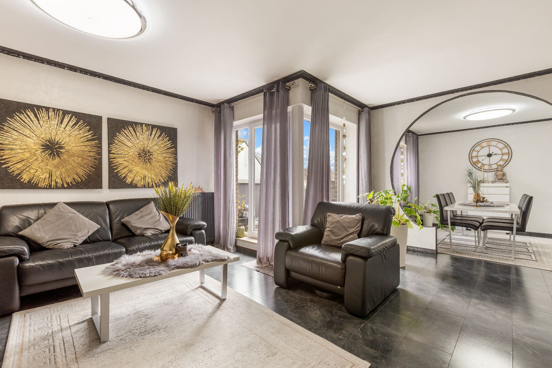 Modernes Wohnzimmer mit schwarzem Sofa, Sessel, Couchtisch und Essbereich, große Fenster mit grauen Vorhängen.