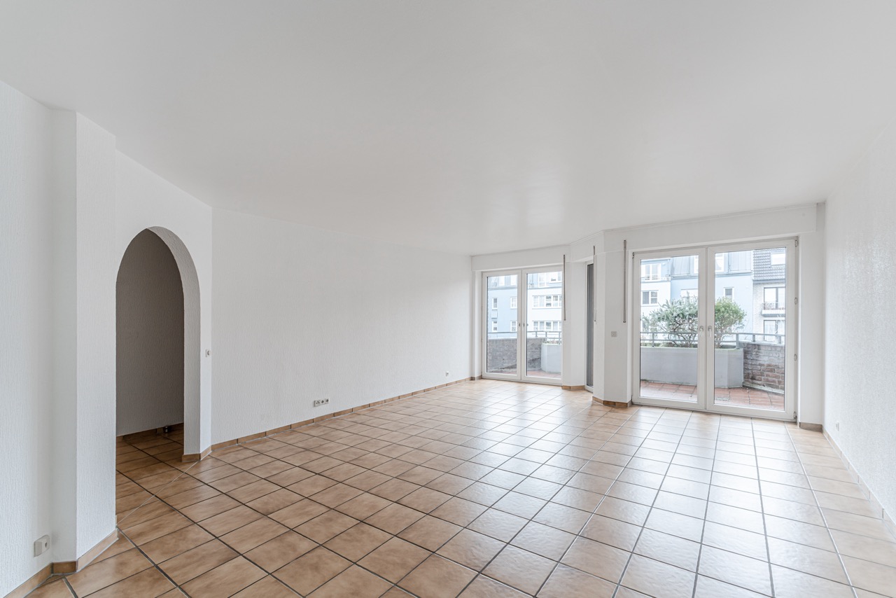 Helles, leeres Wohnzimmer mit Fliesenboden und großen Fenstern zur Terrasse.