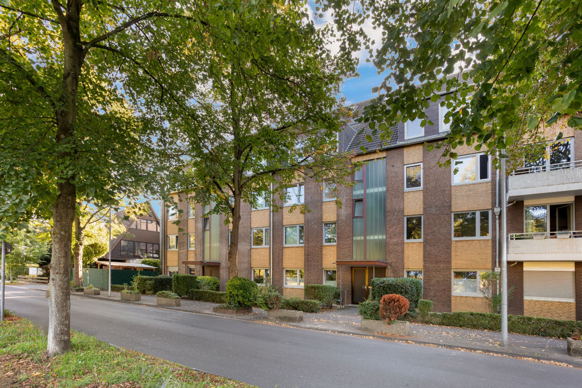 Mehrfamilienhaus mit Klinkerfassade und großen Fenstern an einer baumbestandenen Straße.