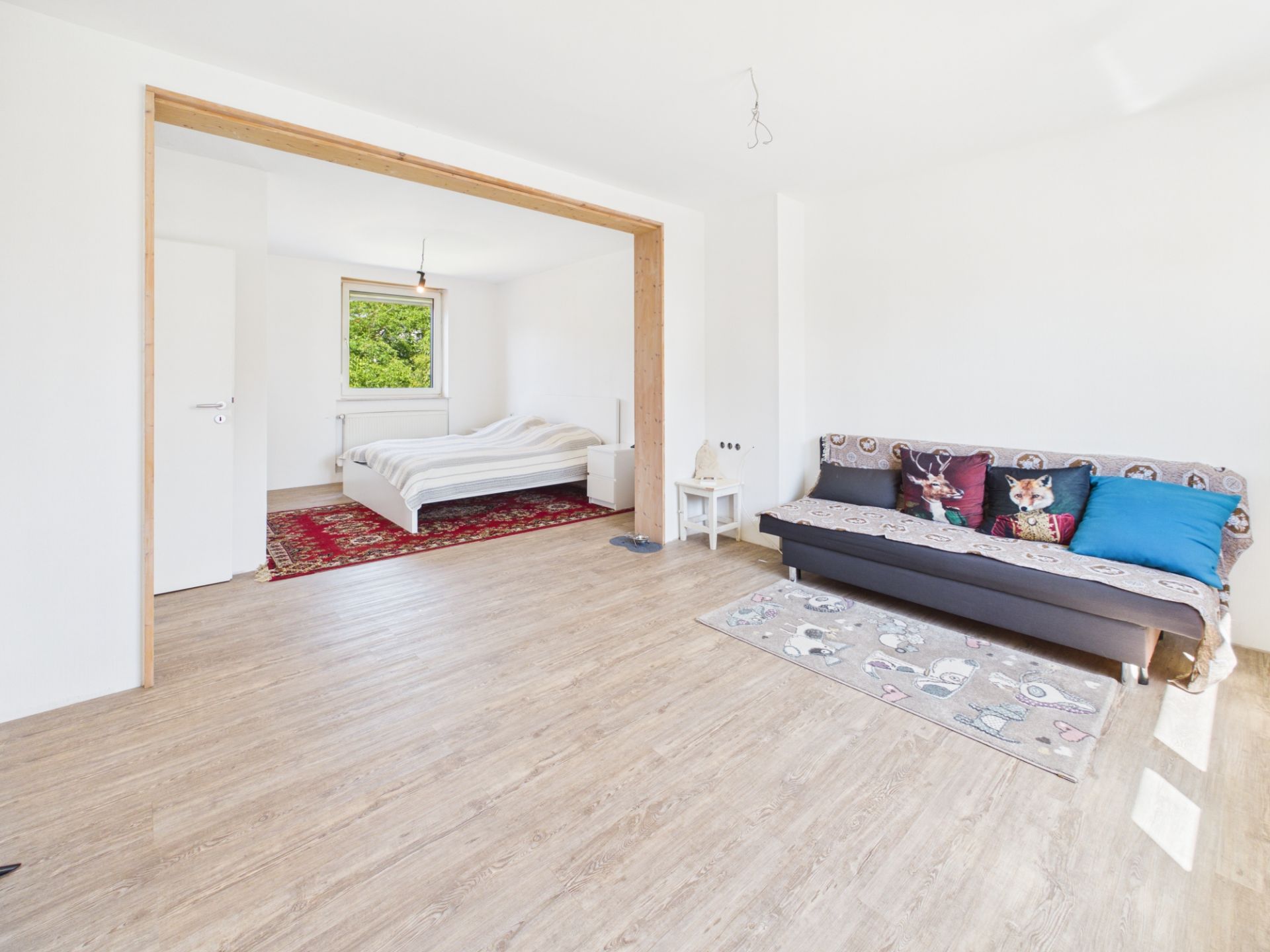 Helles Zimmer mit Holzboden, Schlafbereich mit Bett und Teppich, Couch mit Kissen im Vordergrund.