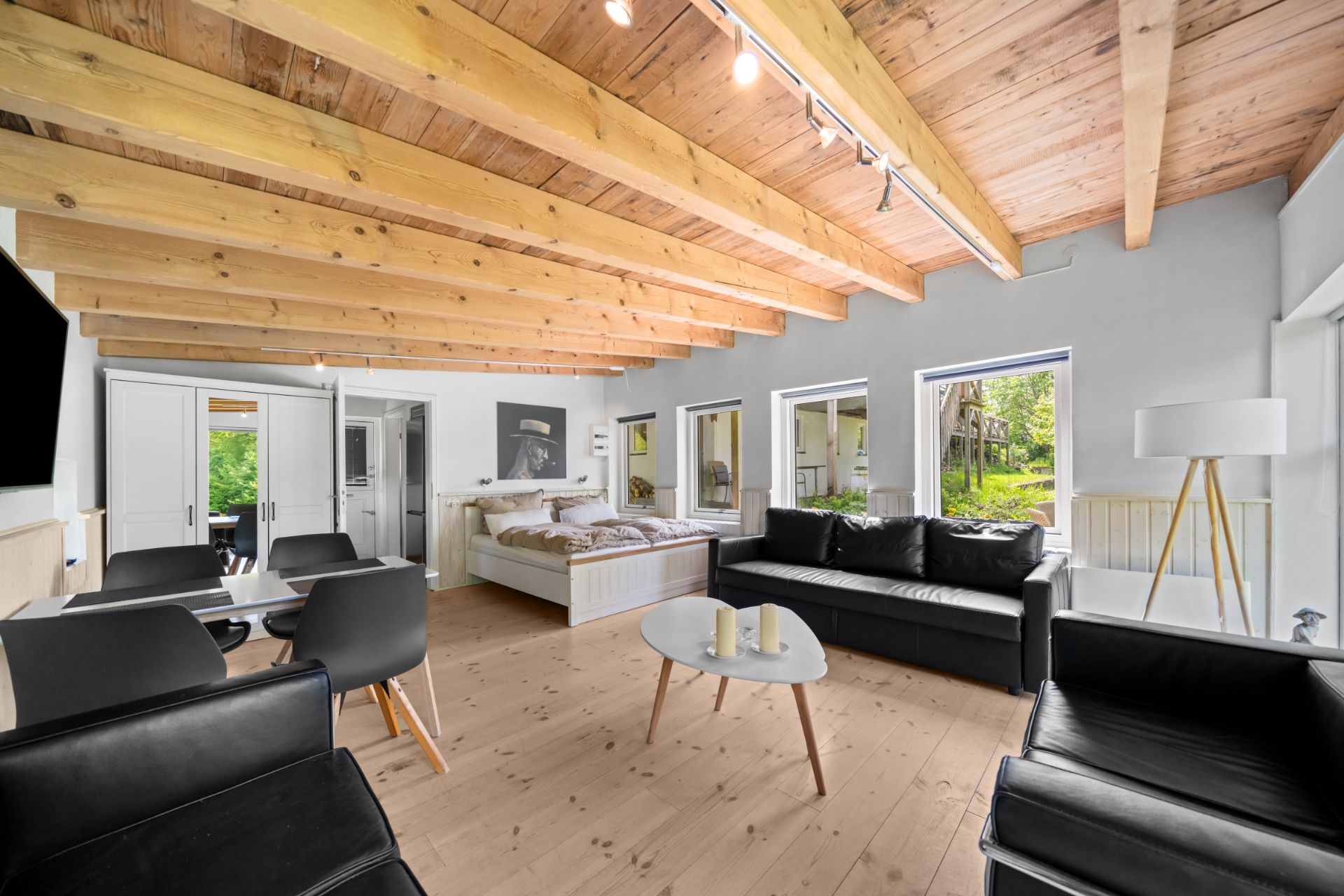 Helles Wohnzimmer mit Holzbalkendecke, schwarzen Sofas, Esstisch und Bett, große Fenster mit Blick ins Grüne.