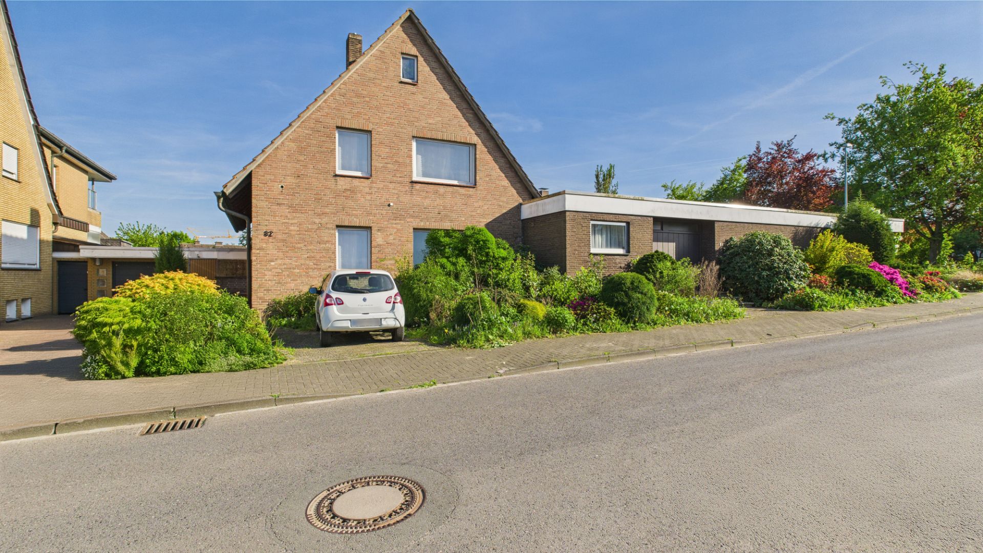 Haus aus Backstein mit Garten und geparktem Auto an einer Straße, umgeben von Büschen und Blumen.
