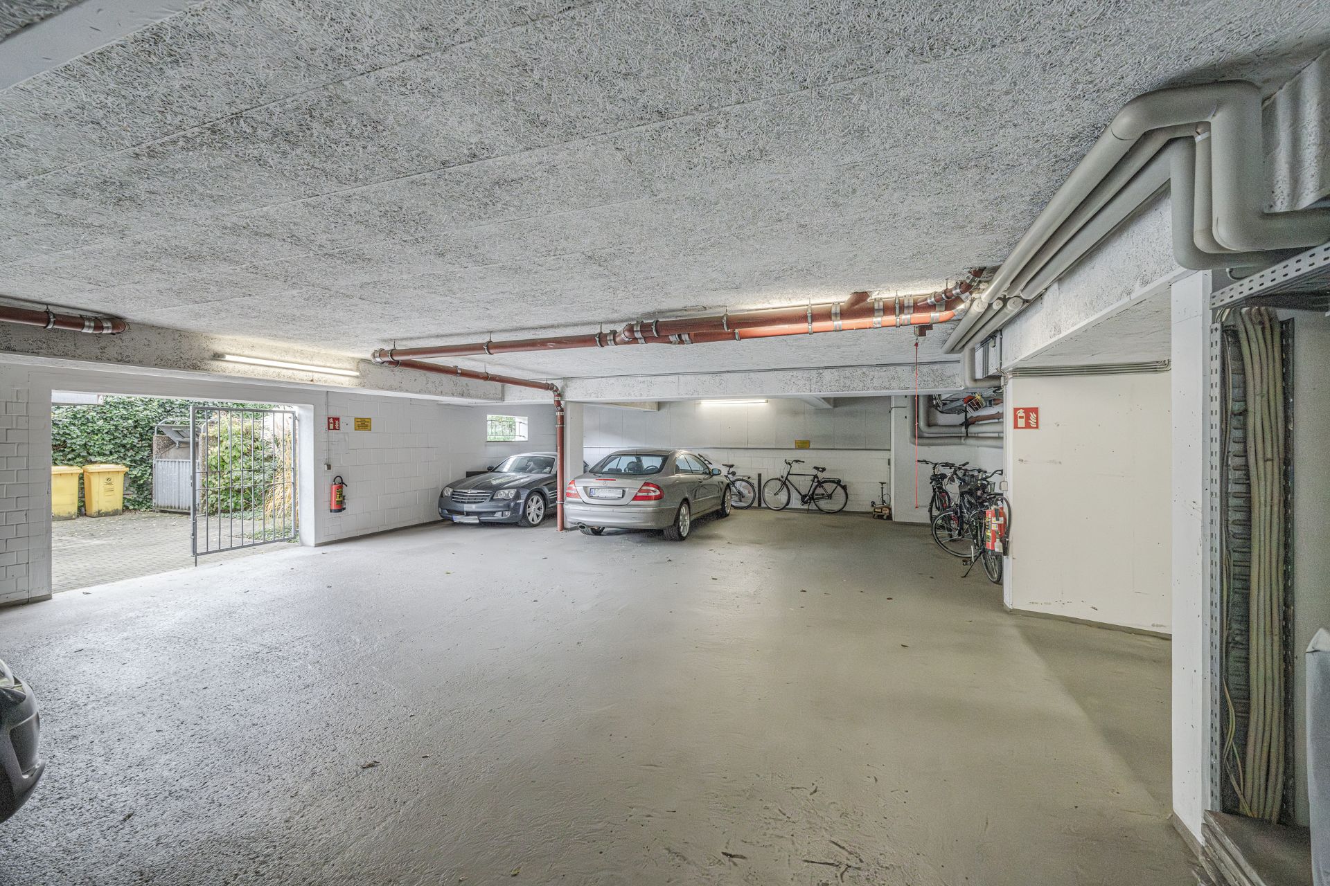 Helle Tiefgarage mit geparkten Autos und Fahrrädern, offenem Tor und sichtbaren Rohren an der Decke.